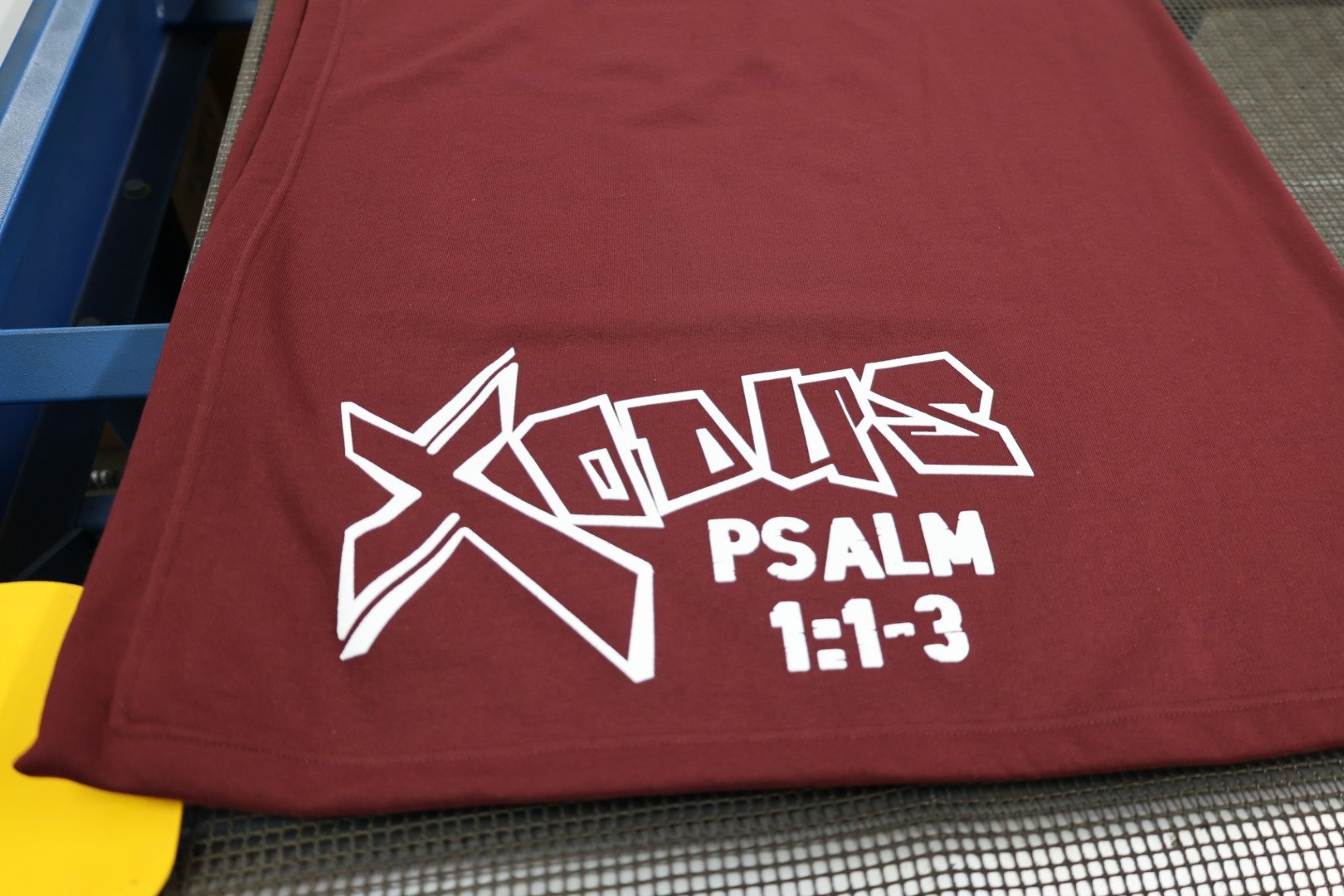 A maroon fabric with white text that reads 'XGLOW PSALM 1:1-3' and a stylized star logo, lying on a black mesh surface with part of a blue frame visible in the background.