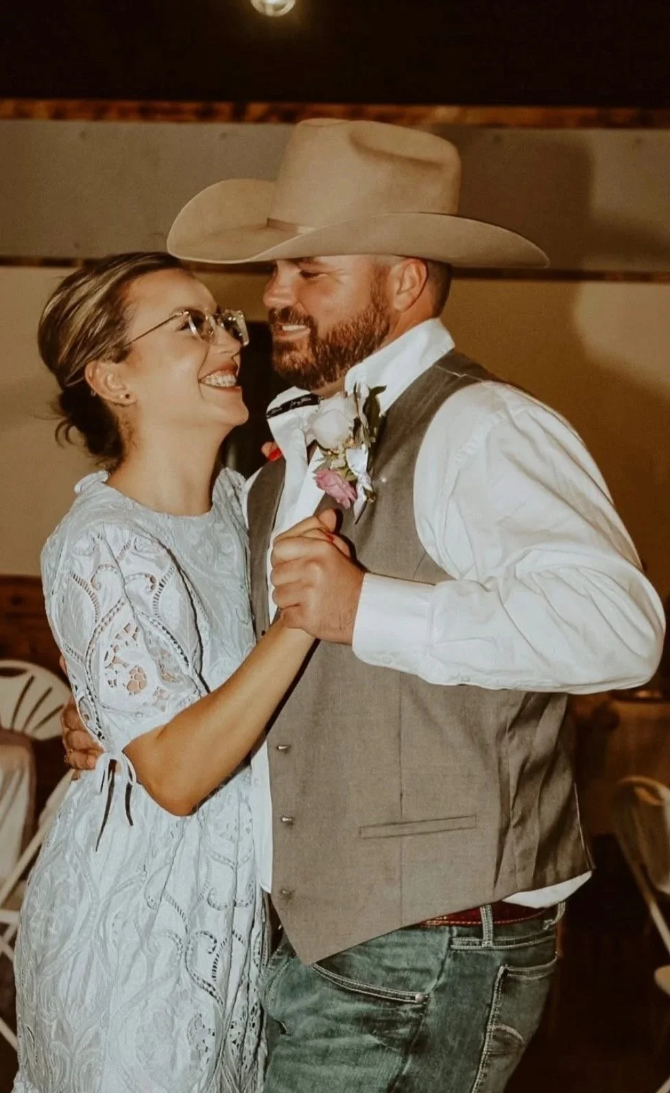 A couple music and cowboy-themed wedding dance, smiling and looking at each other, with the woman in a white lace dress and glasses, and the man in a cowboy hat, vest, and jeans.