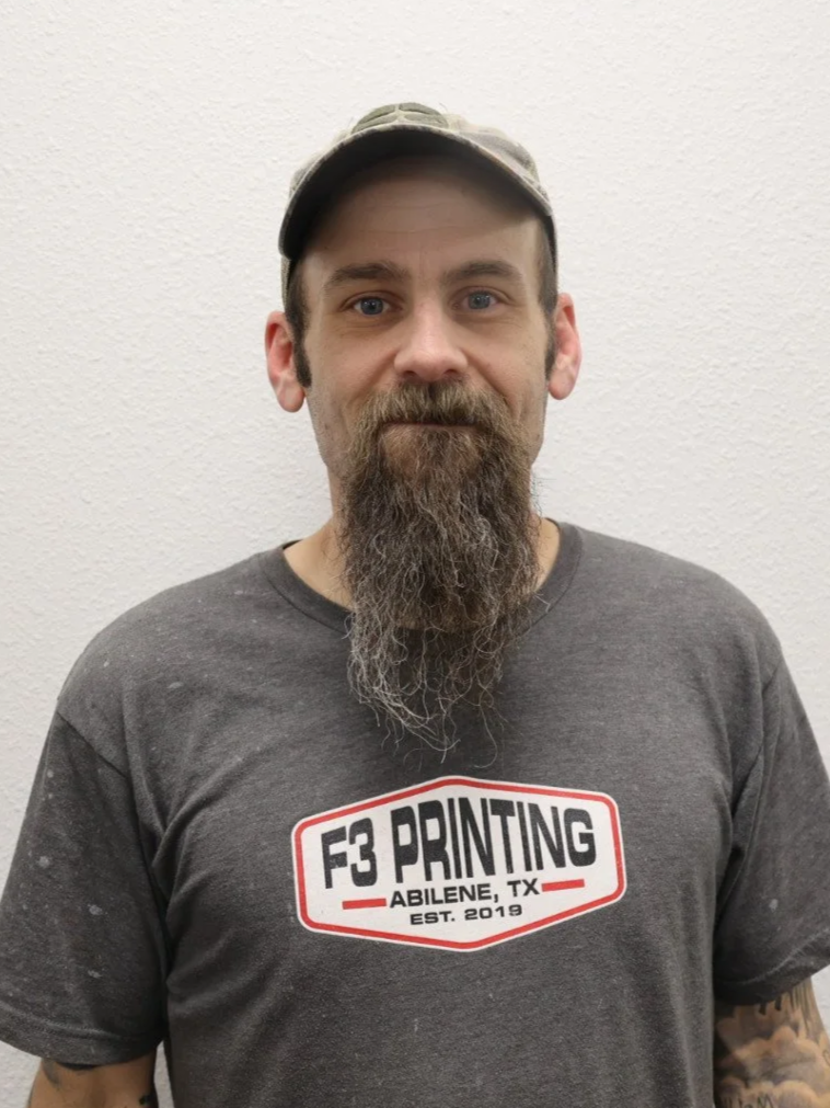 A man with a beard and mustache wearing a camouflage cap and a dark gray T-shirt with a logo for F3 Printing in Abilene, Texas.