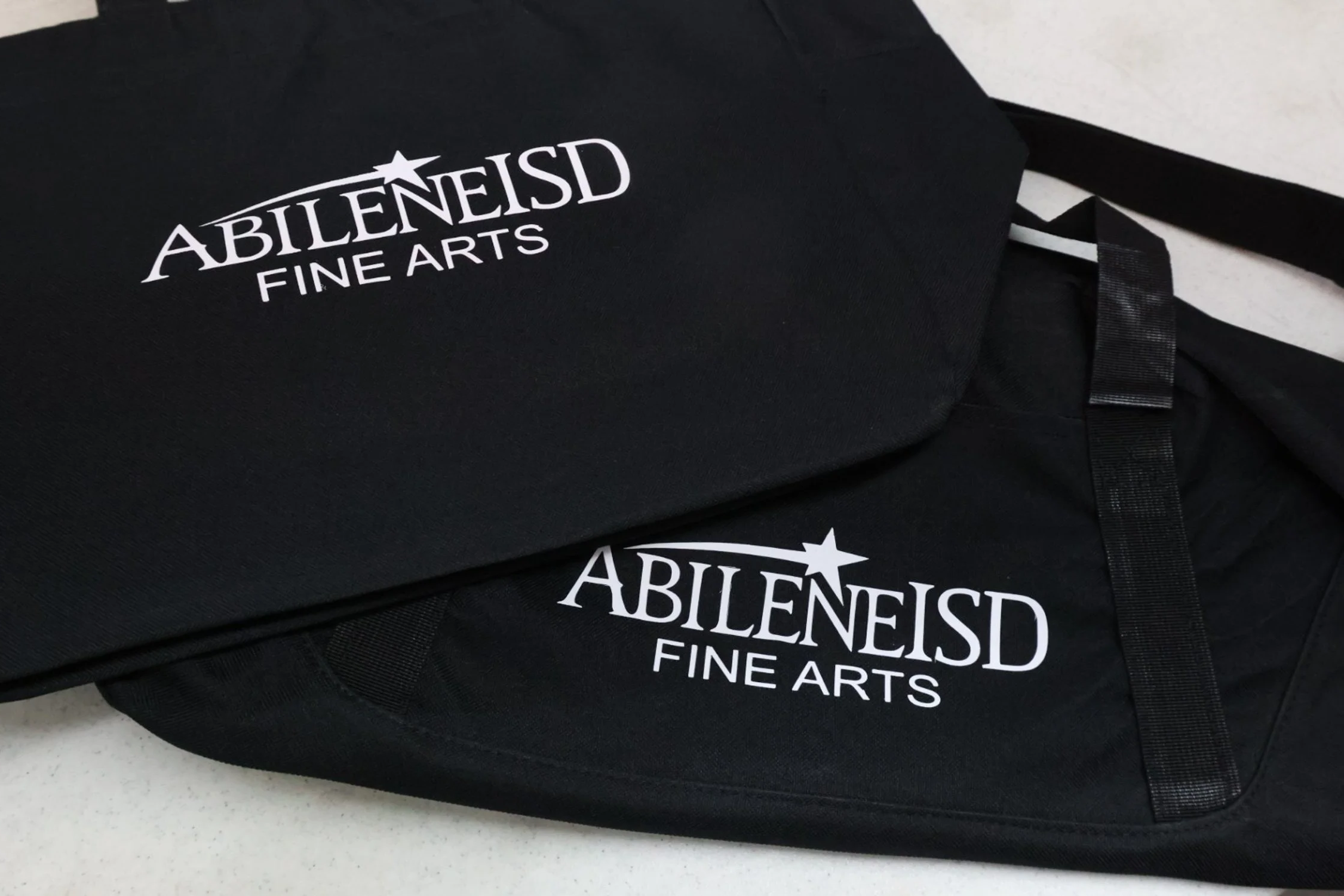 Two black bags with white logo text that reads 'ABILENE ISD FINE ARTS' placed on a light-colored surface.