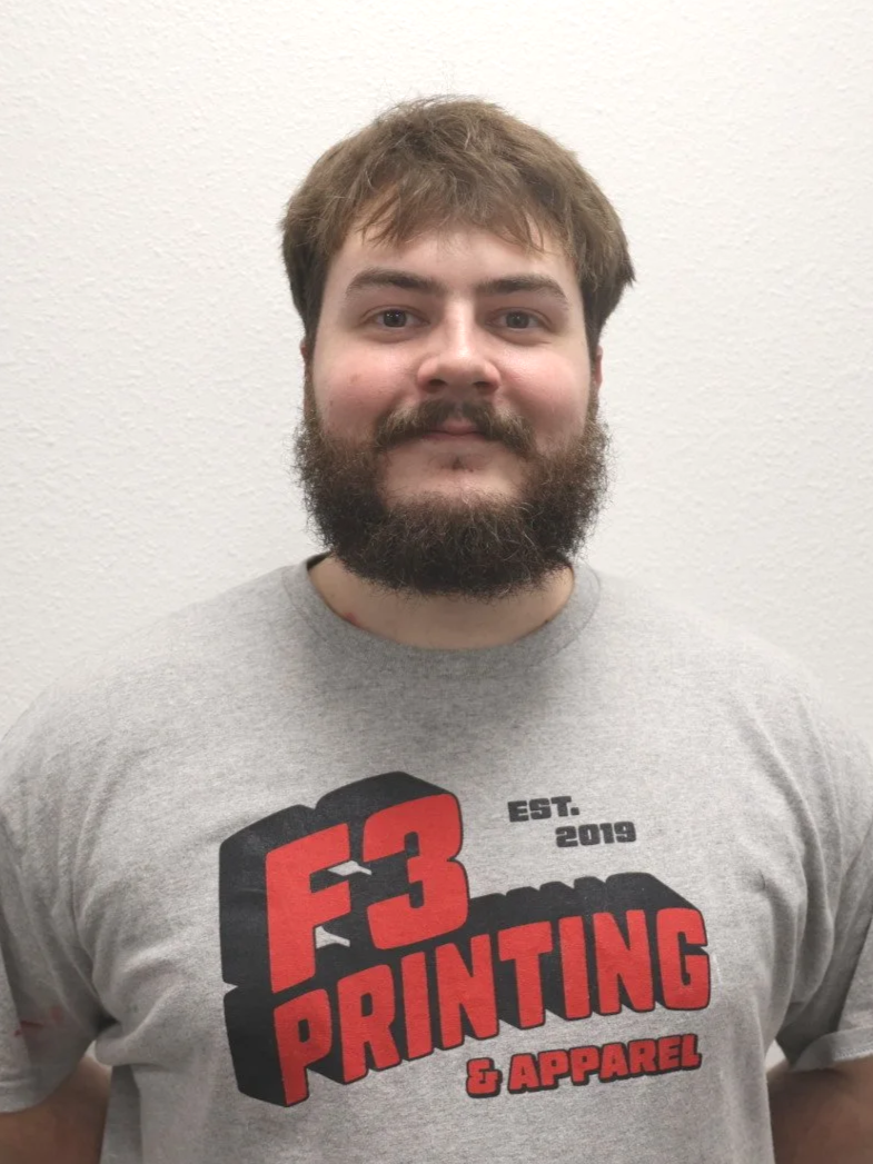 A young man with brown hair, a beard, and mustache wearing a gray T-shirt with a logo that says "F3 Printing & Apparel," standing against a plain white wall.