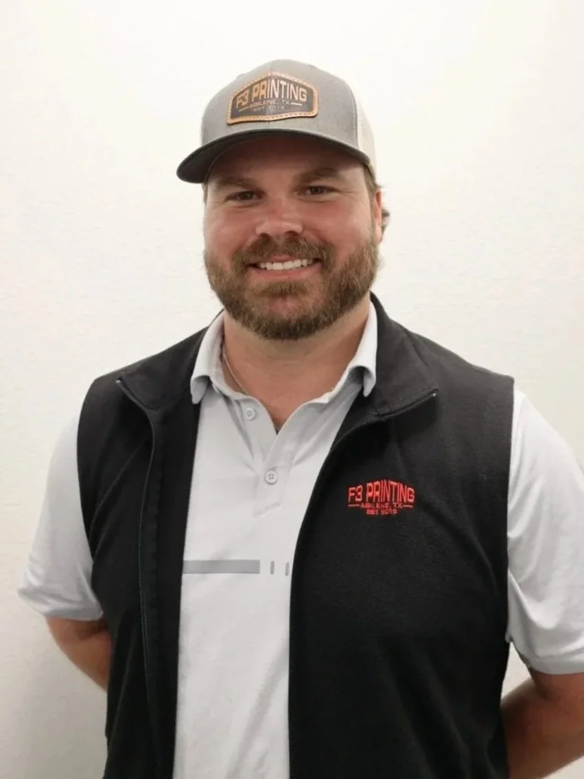 A smiling man wearing a gray cap with a logo, a white polo shirt, and a black vest with a logo, standing against a plain white wall.