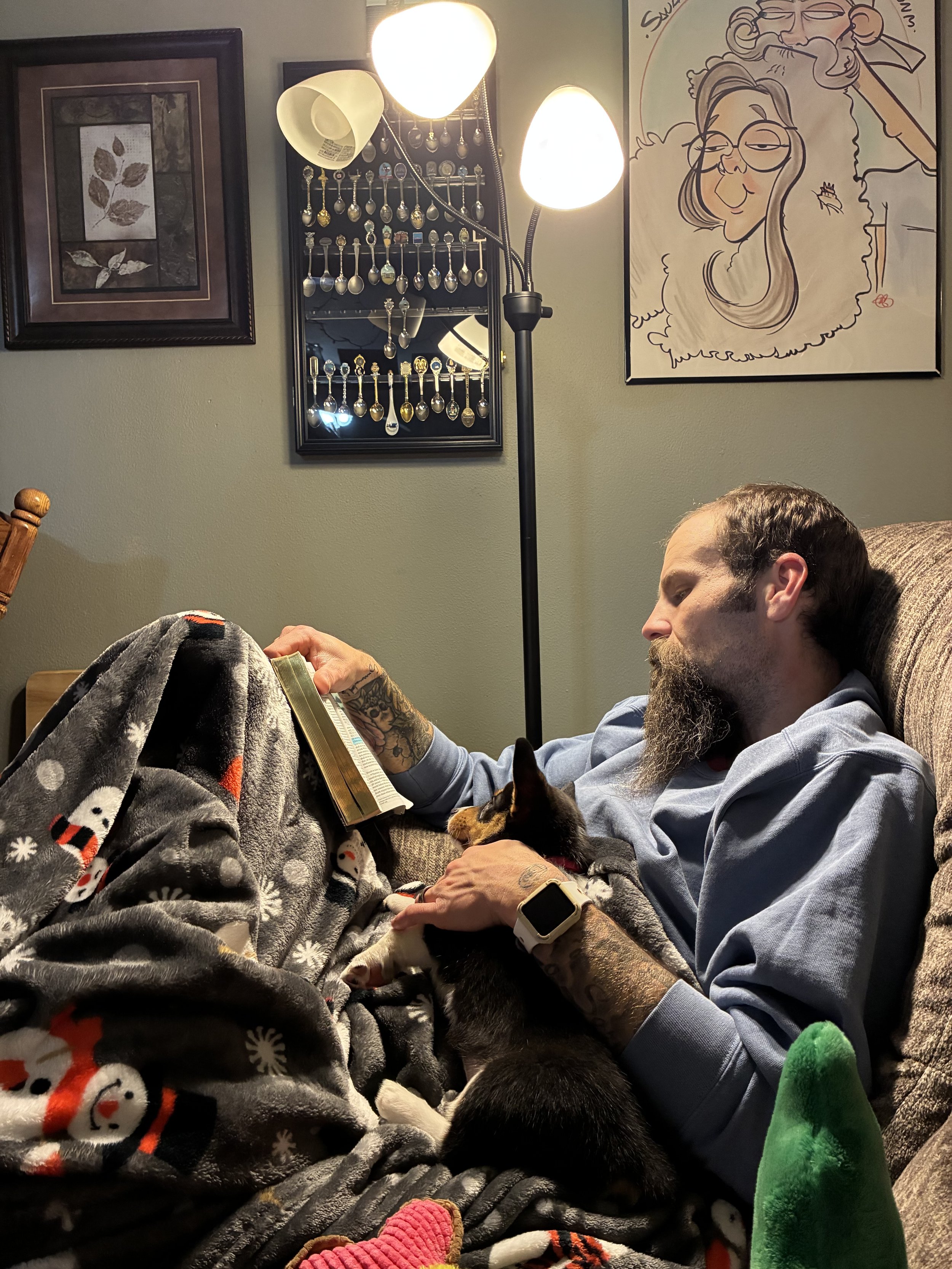 A man with a beard and tattoos is sitting on a couch, reading a book with a cat on his lap. There are multiple lamps and framed artwork on the wall behind him, including a large caricature drawing of a woman with glasses and a man. The man has a smartwatch on his wrist and is wearing a light blue shirt.
