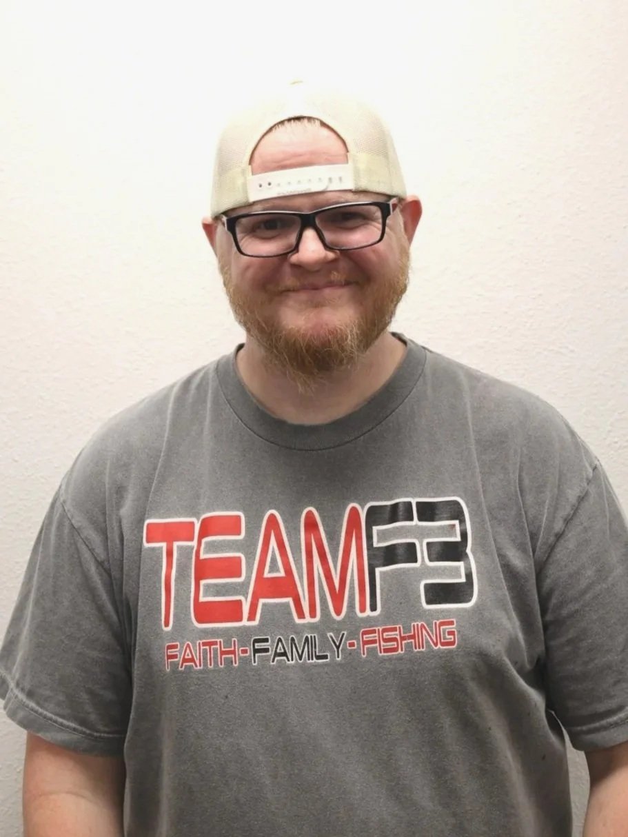 A man with glasses and a beard wearing a gray ''TEAM F3'' T-shirt that says ''FAITH - FAMILY - FISHING'' and a backwards beige baseball cap, smiling, standing against a plain white wall.