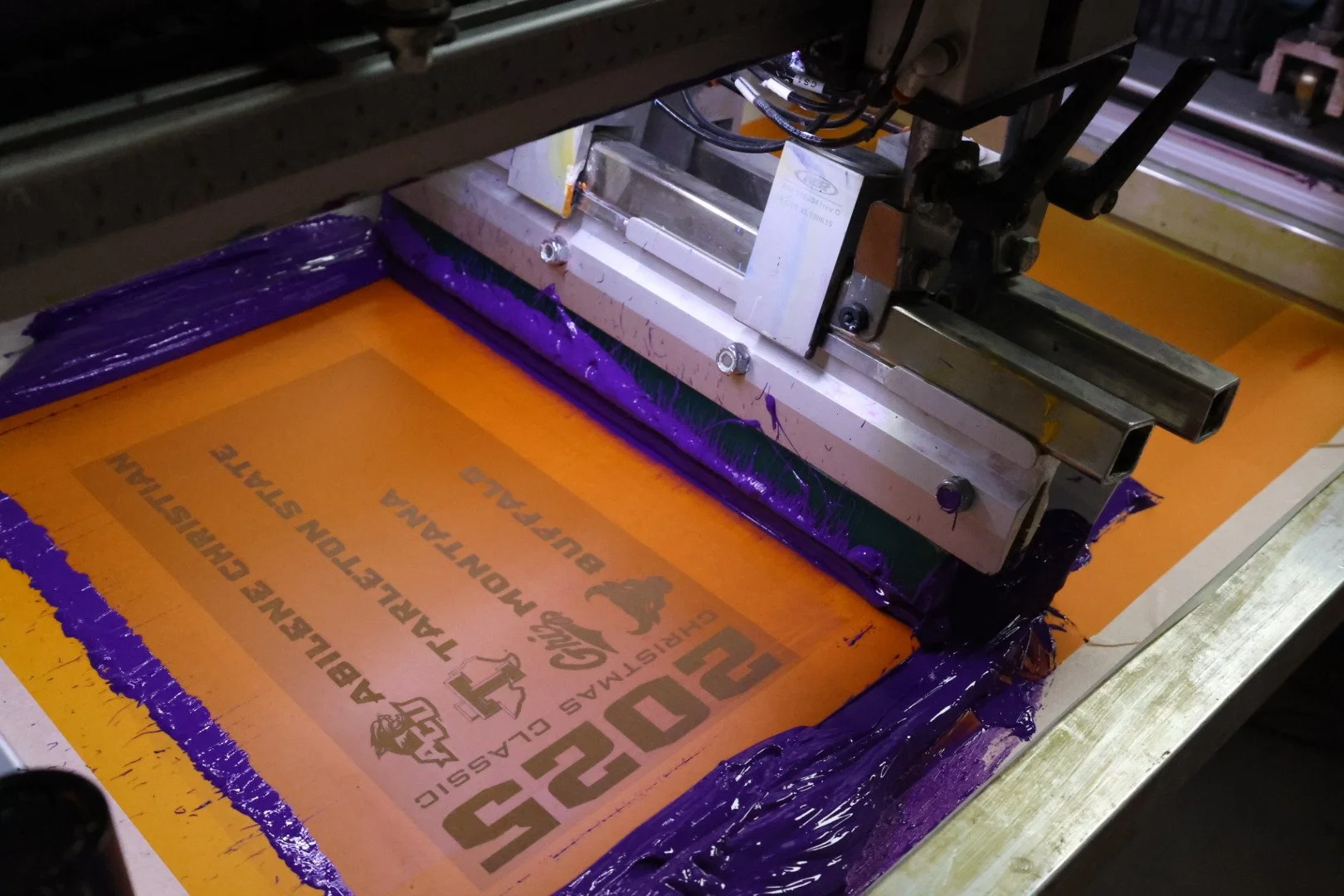 Screen printing machine printing on orange and purple fabric with text and logos.