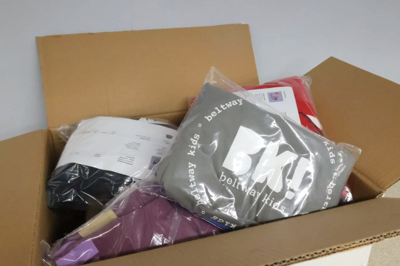 Open cardboard box containing packaged clothing and supplies, including a gray clothing item labeled 'beltway kids' in a plastic bag.