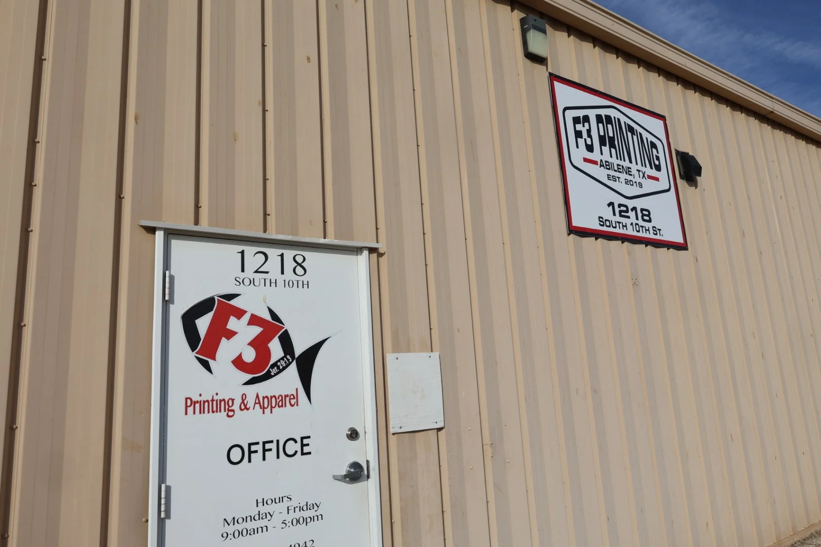 Exterior view of a beige metal building with signage for F3 Printing, including a sign on the wall and a door sign indicating the office address, hours, and services, located at 1218 South 10th Street, Abilene, TX.