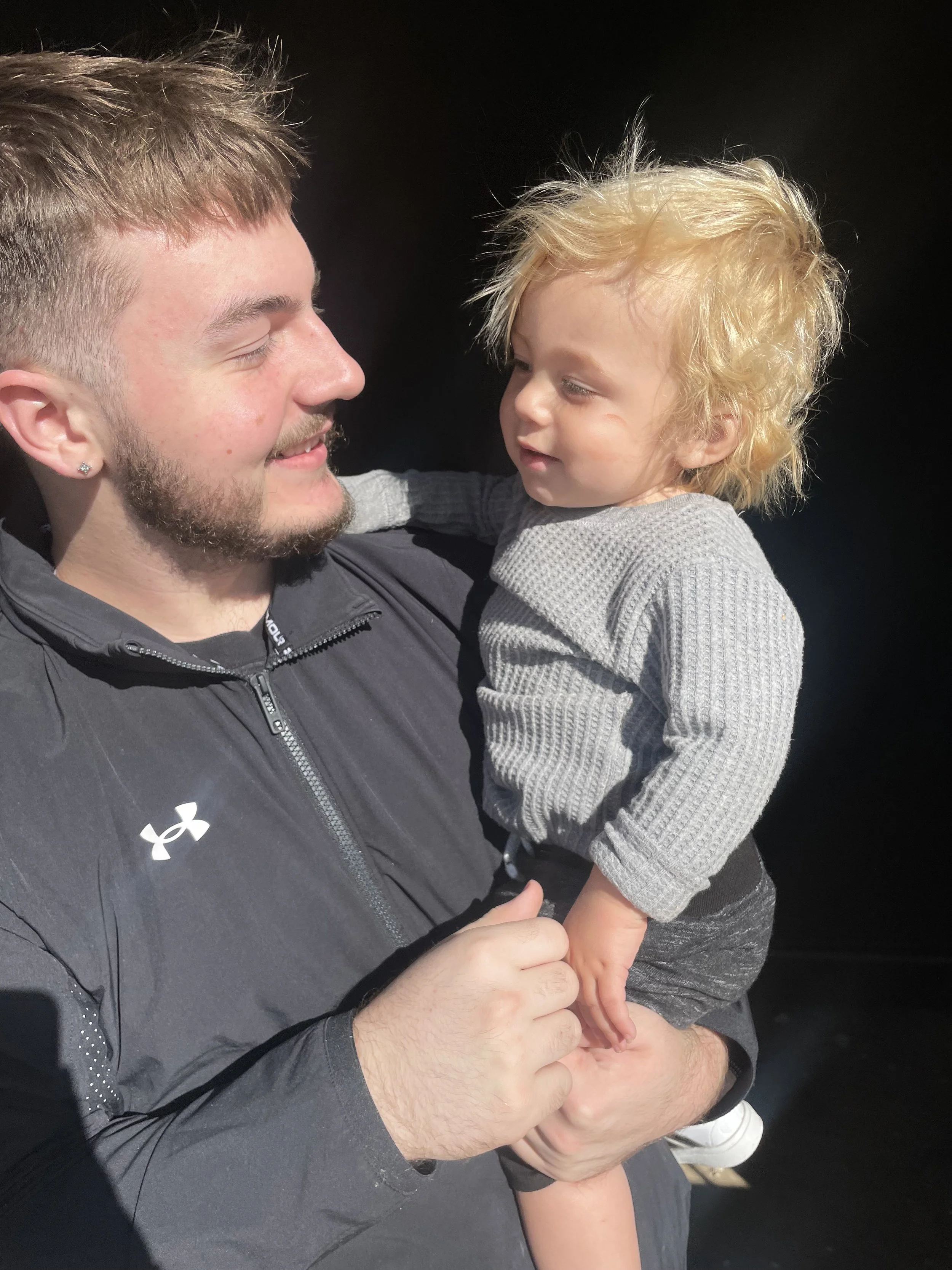 A man with short light brown hair, a beard, and wearing a black jacket is holding a young child with curly blonde hair and a gray sweater, against a black background.