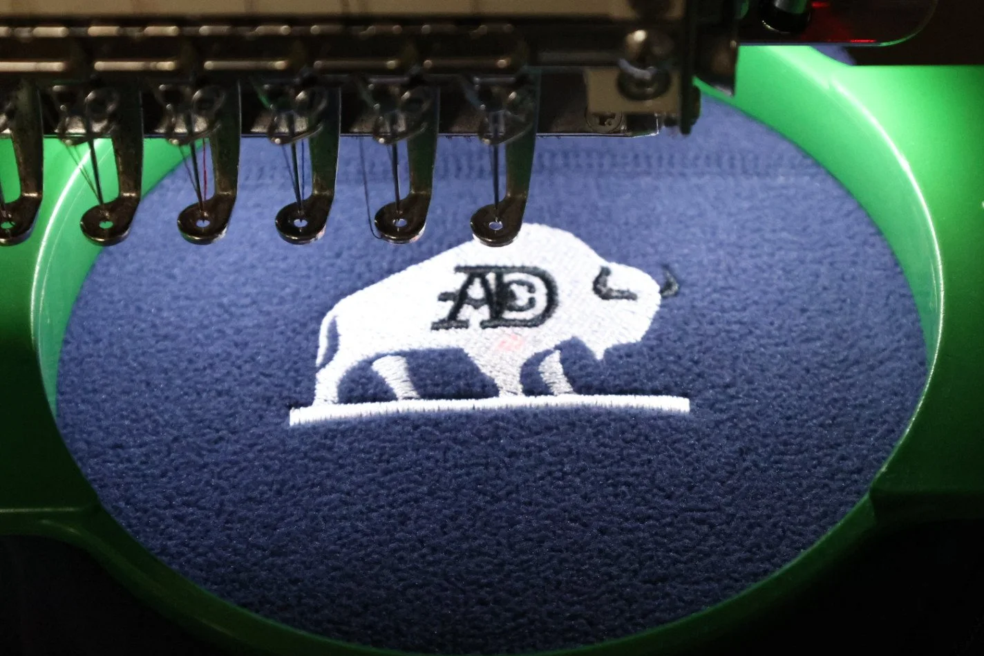 Embroidery machine stitching a patch with a panda bear design and the initials 'A D' on a blue fabric.