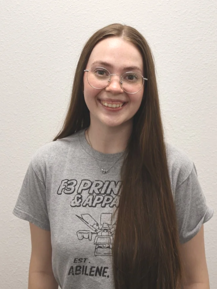 A young woman with long brown hair, glasses, and a gray T-shirt, smiling and standing against a plain white wall.