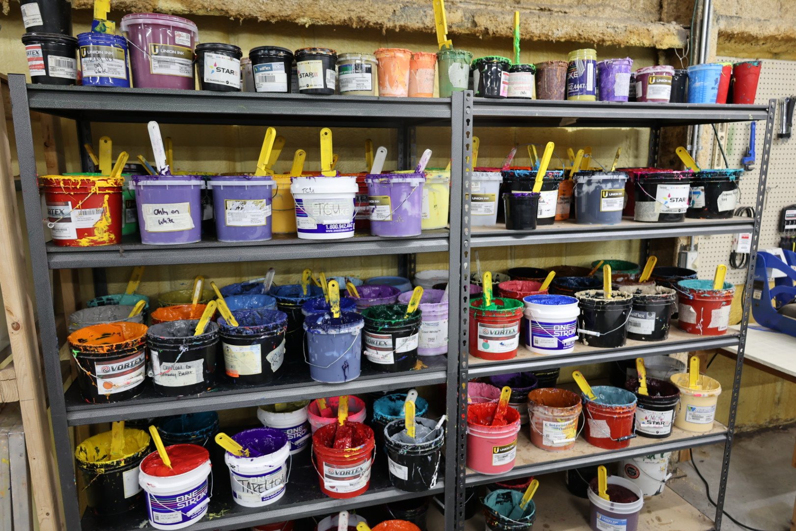 Metal shelving unit filled with paint buckets in various colors, with some buckets having paint drips and stains around the rims. The buckets have paint mixing sticks sticking out of them, and some have handwritten labels.