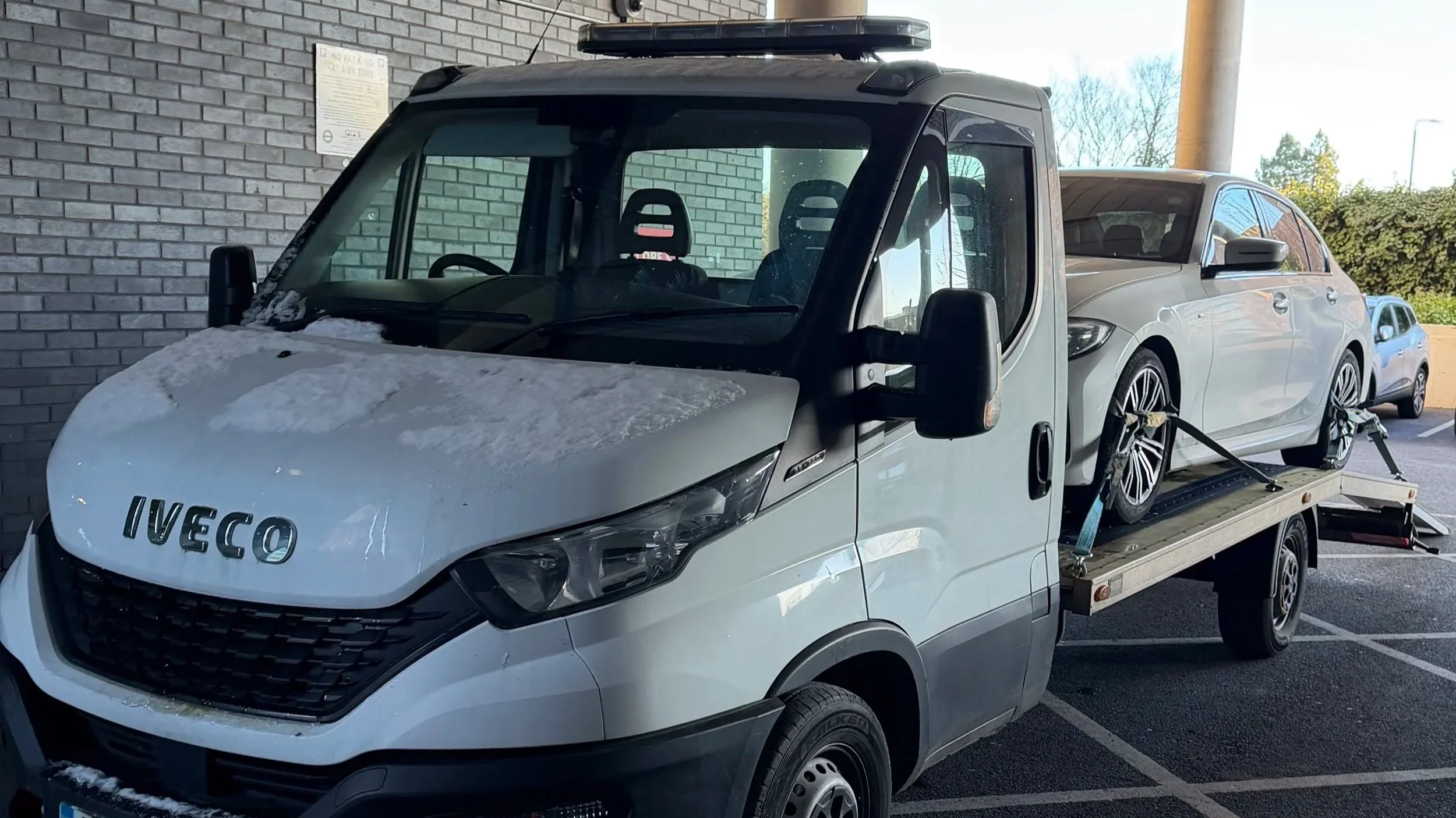 A white Iveco flatbed tow truck transporting a silver hatchback car in a parking lot, with other cars and trees in the background.