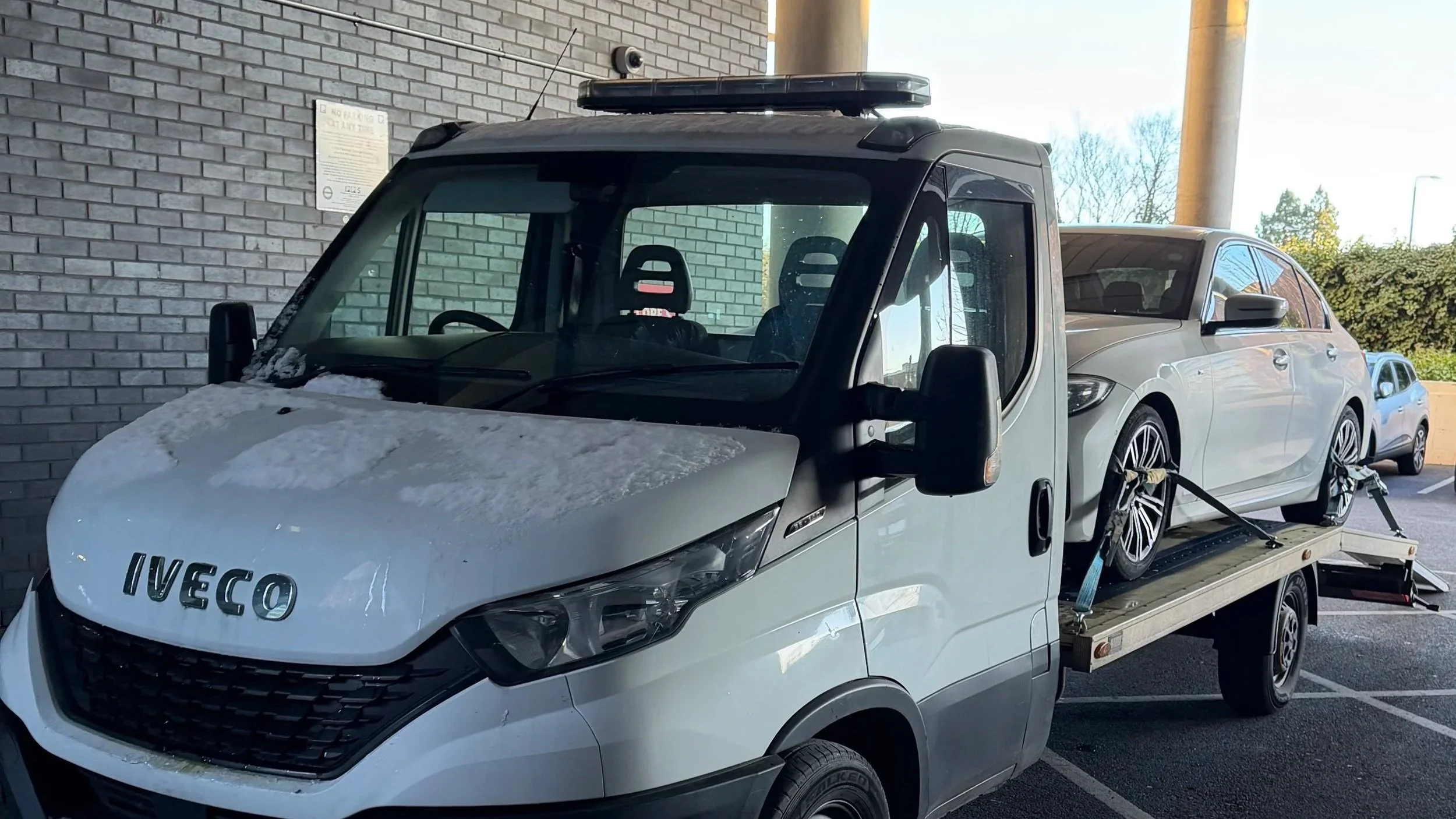 White tow truck with the brand name Iveco towing a silver hatchback car in a parking lot.