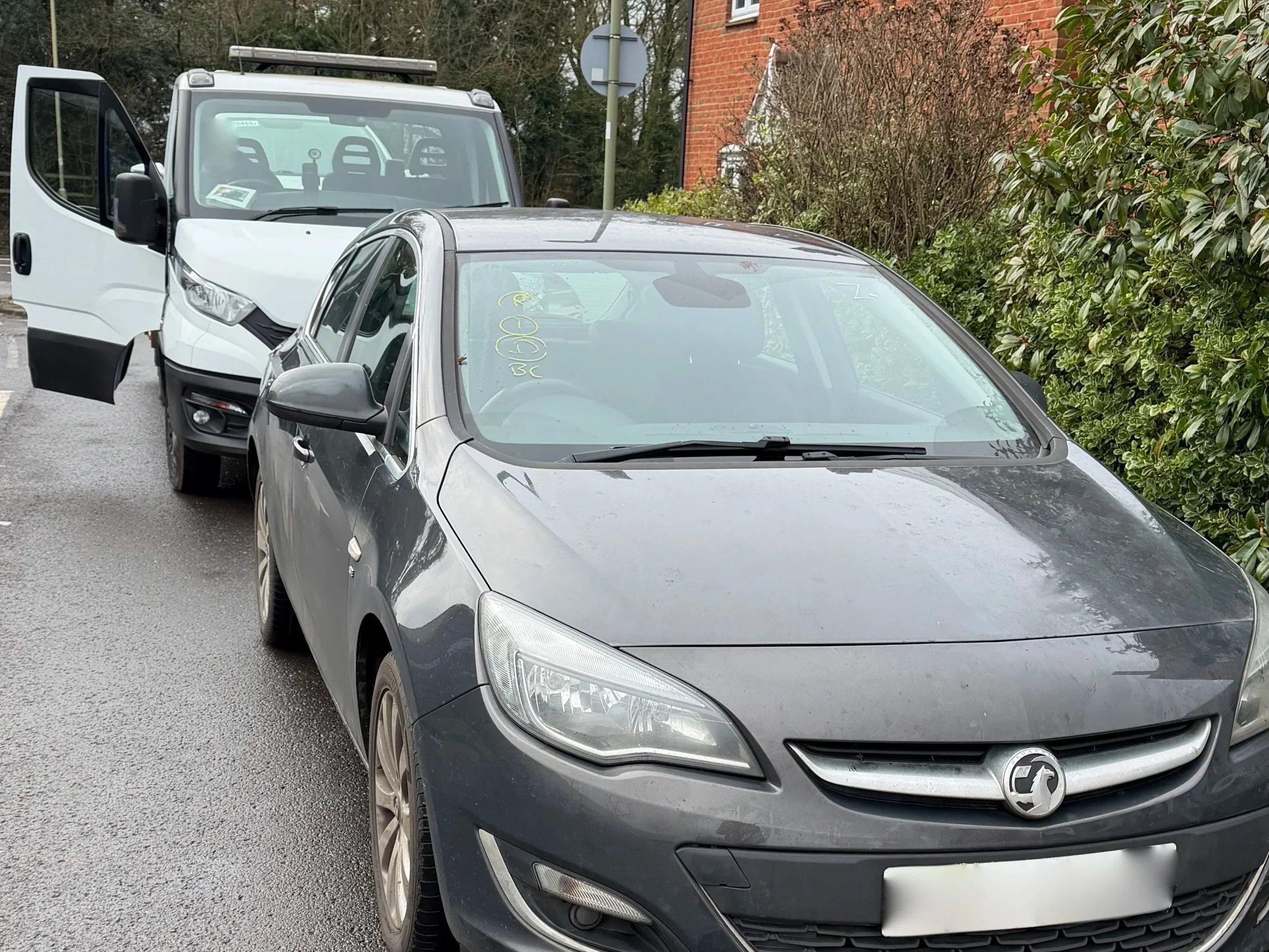 Gray Vauxhall car parked on the side of the street with a white van behind it, next to a green hedge and a brick building
