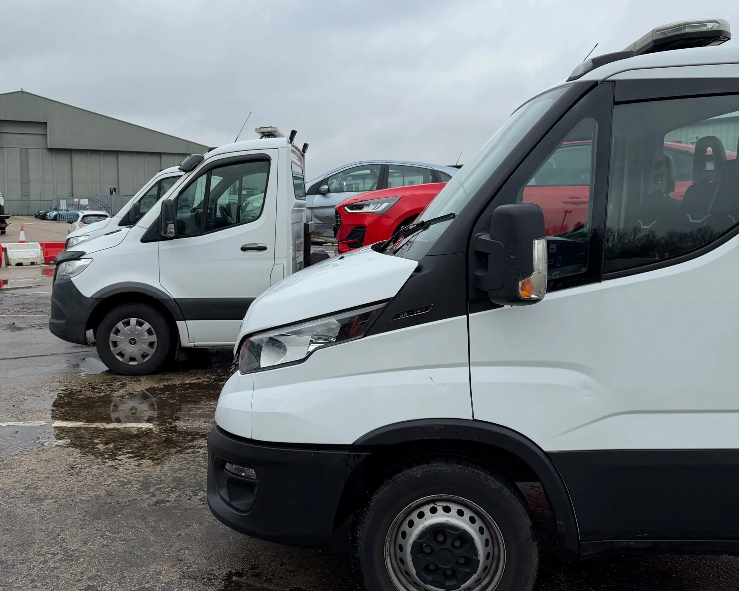 Multiple white commercial vans parked outdoors on a cloudy day, with a red car in the background.