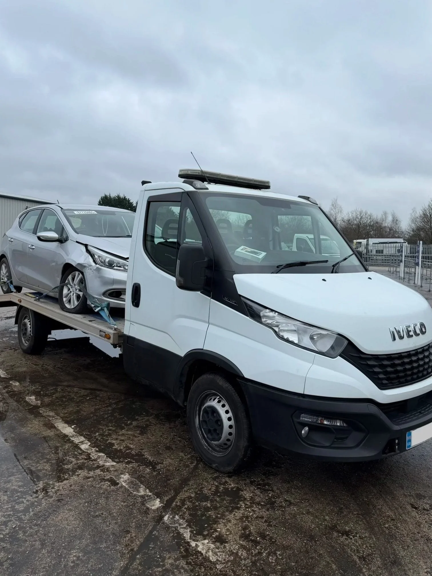 A white flatbed tow truck carrying a silver damaged car with front-end damage, parked on a wet outdoor lot under a cloudy sky.