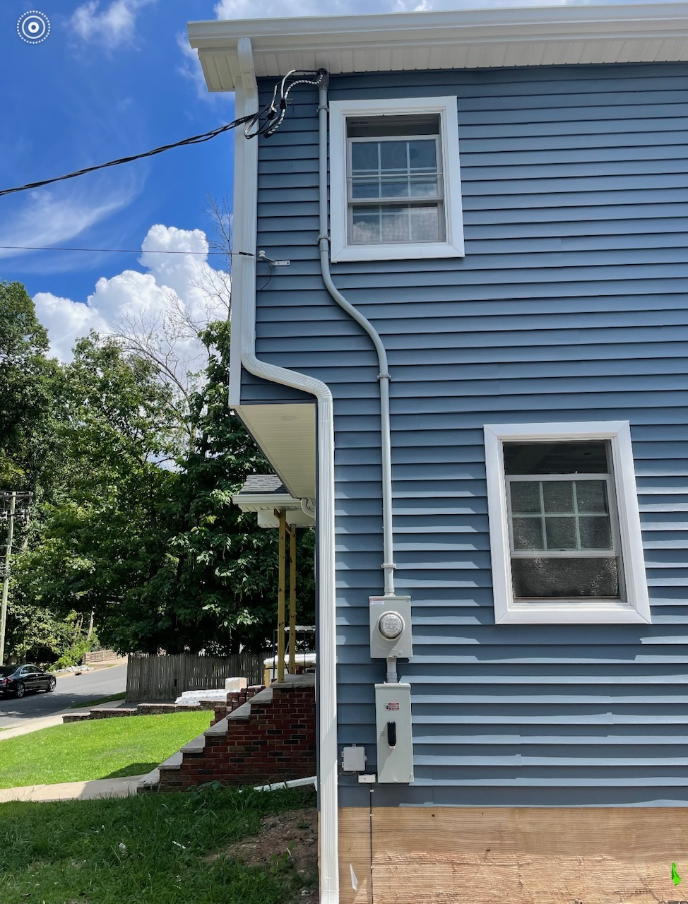 Exterior of a blue house with two white-framed windows, electrical wiring, and a utility box on the side wall, with stairs and a sidewalk nearby, under a partly cloudy sky.