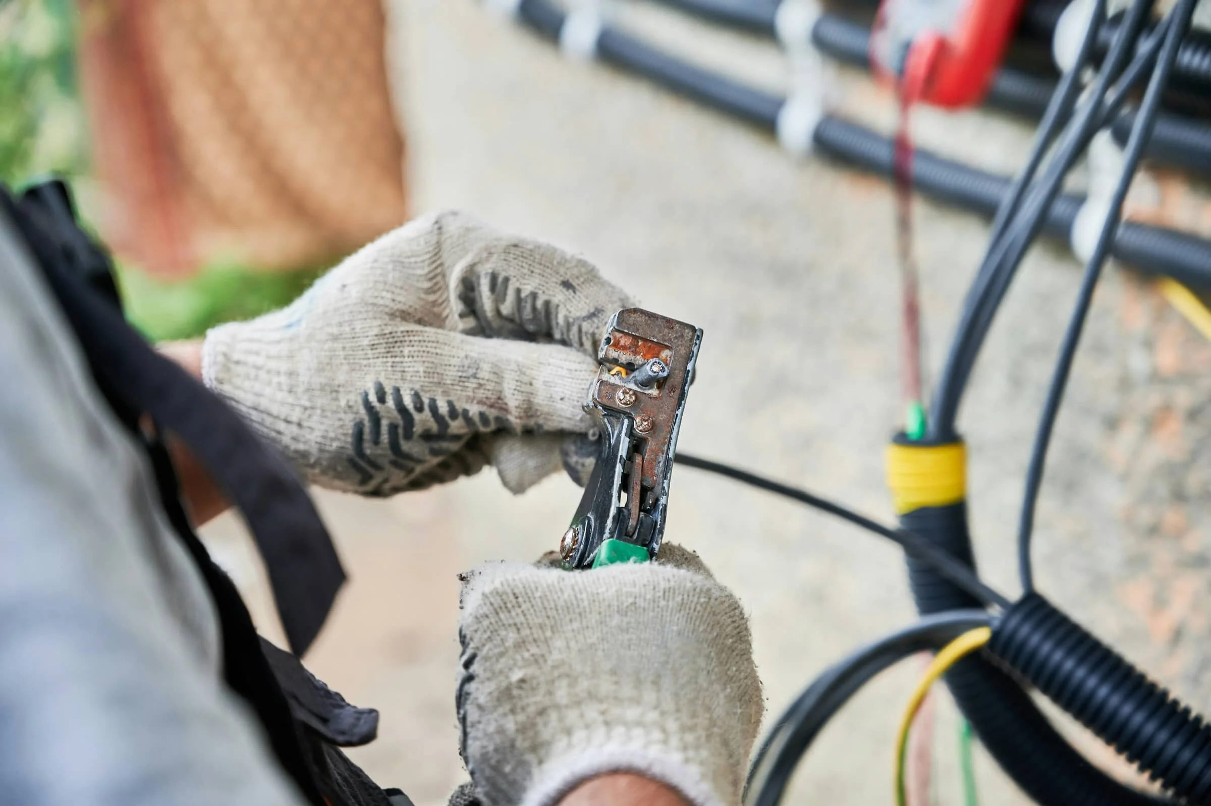 Close-up of worker's hands wearing gloves, using pliers to work on electrical wiring with multiple cables and tools visible in the background.