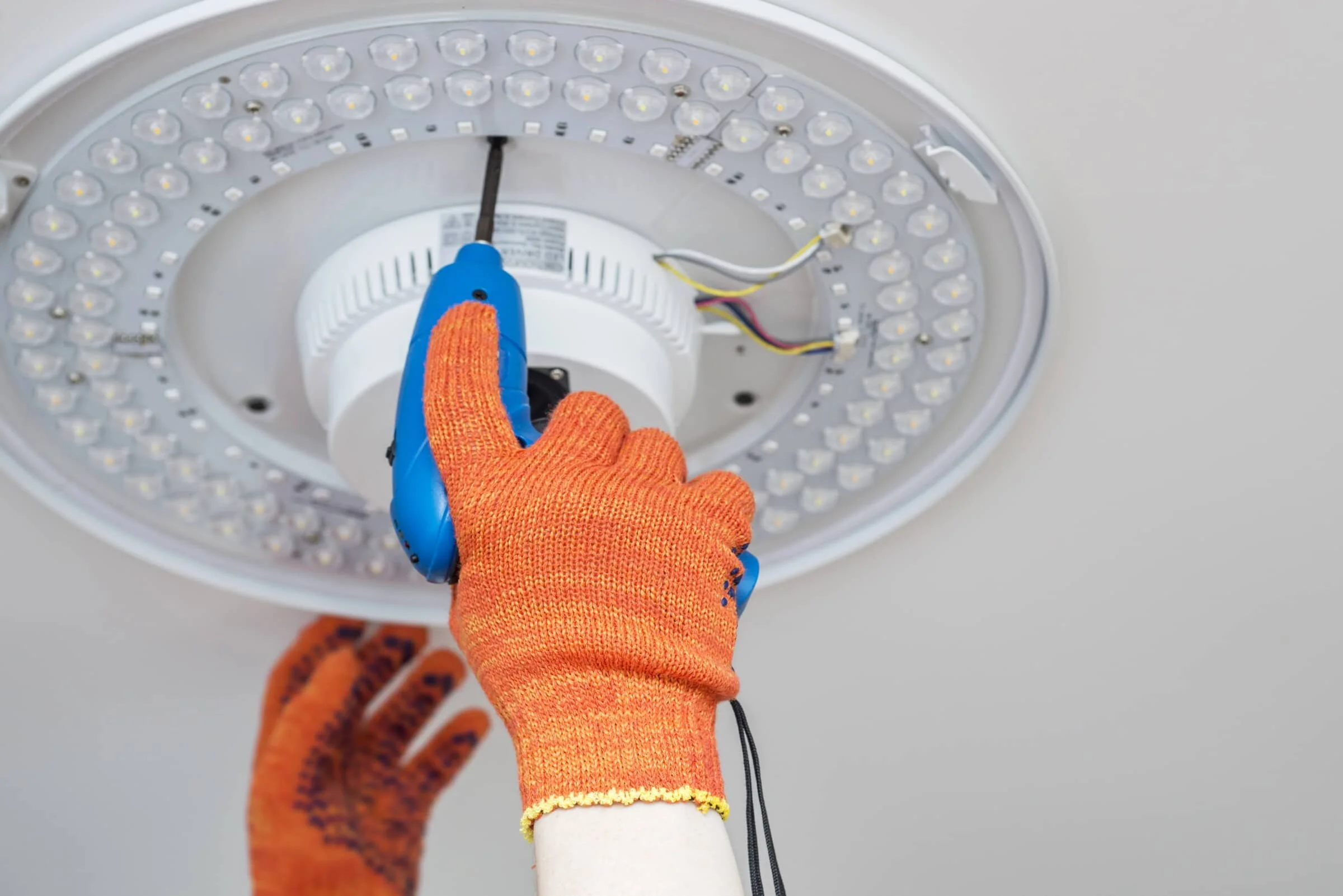 Person using a blue cordless drill to work on a circular LED ceiling light fixture, wearing orange gloves.