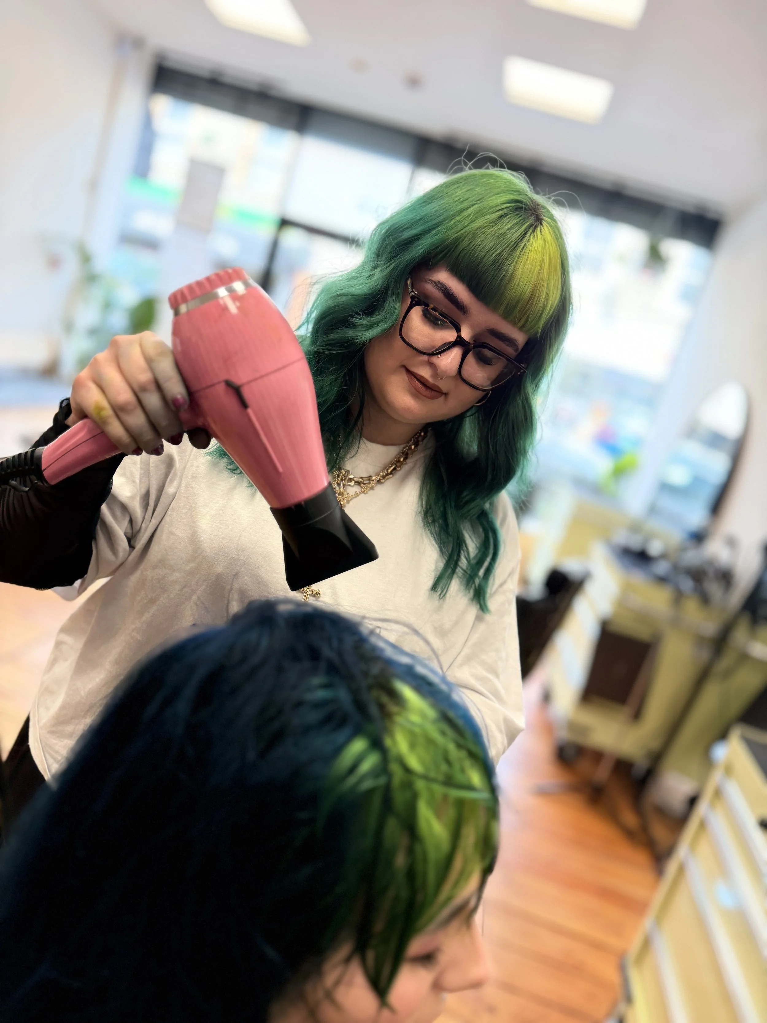 Woman with green hair styling a customer's hair in a salon.