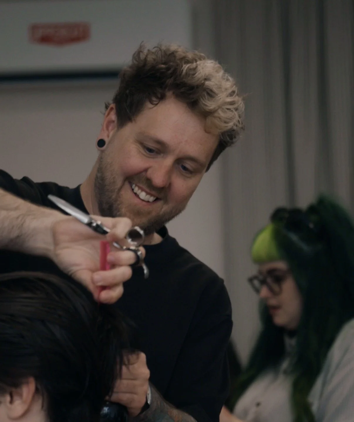 A smiling man with curly hair is giving a haircut to a customer with dark hair.