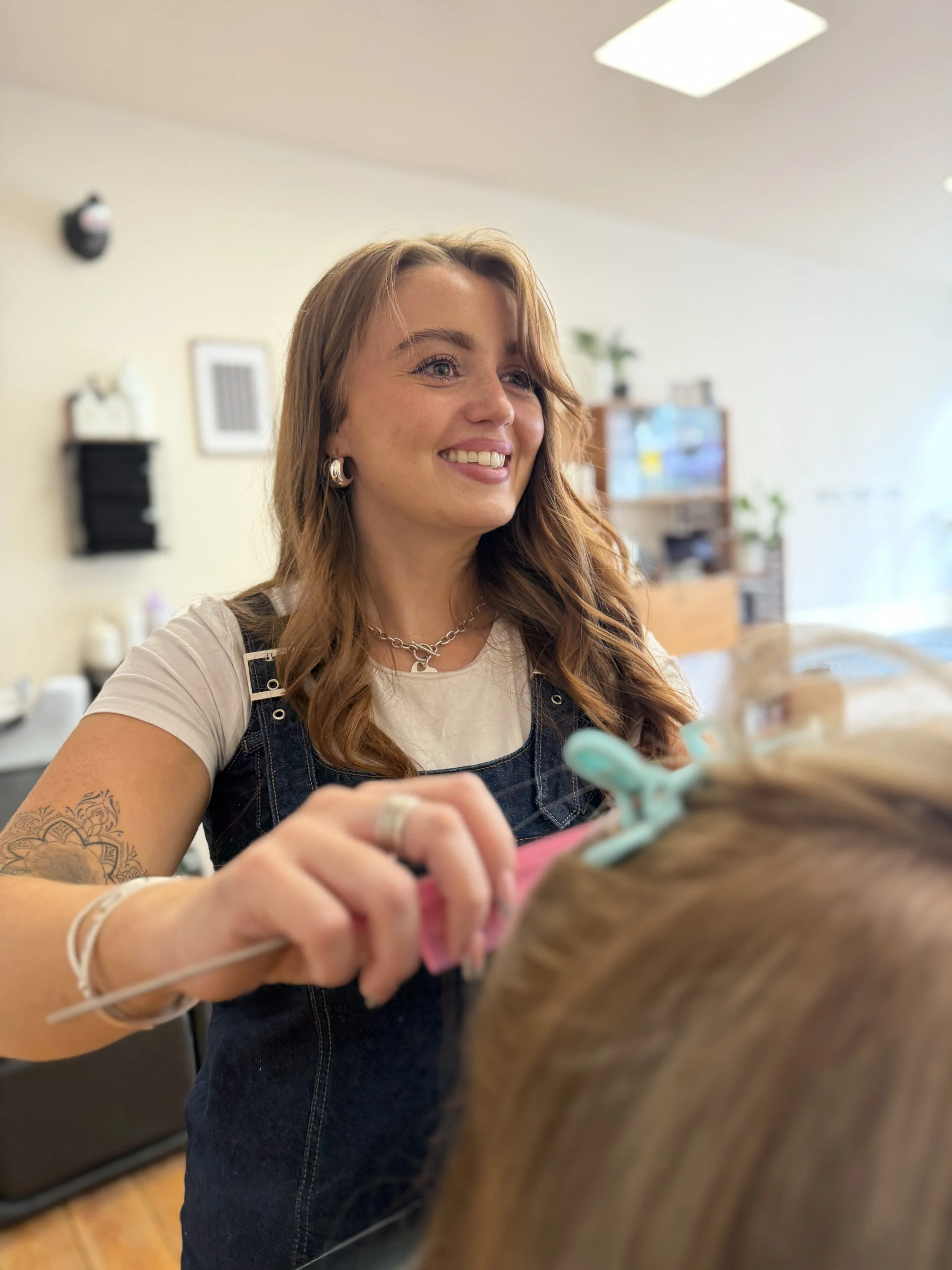 A woman with long wavy hair, wearing a white shirt, denim overalls, earrings, and necklaces cutting a clients hair.