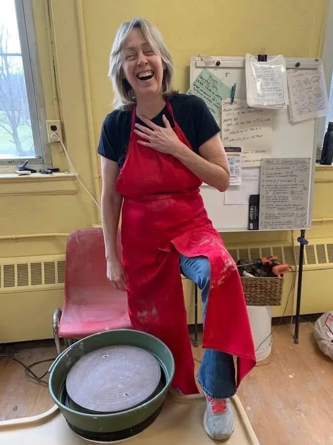 Potter Dixie is standing up, laughing, hand at chest, foot up on pottery wheel.