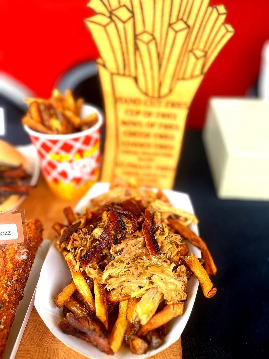A serving of pulled pork with bacon bits and French fries in a white paper boat, with a side of French fries in a red and white checkered container, and a cardboard container of French fries in the background. The food is on a wooden surface.