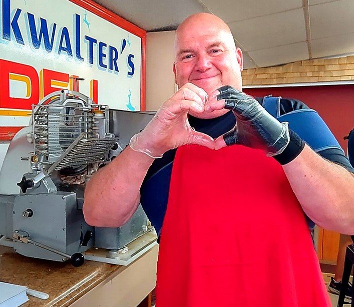 Bucky making a heart shape with his hands in front of a meat slicer, wearing a red apron and a black glove on his right hand, inside a retro-style deli.