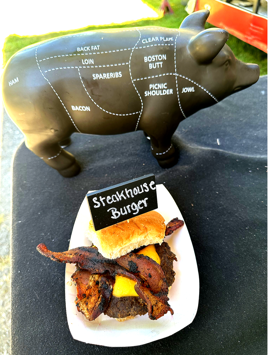 A pig-shaped diagram of a pig roast with labeled cuts of pork, positioned behind a small plate with a bacon cheeseburger labeled 'Steakhous Burger,' featuring bacon, cheese, and a bun.