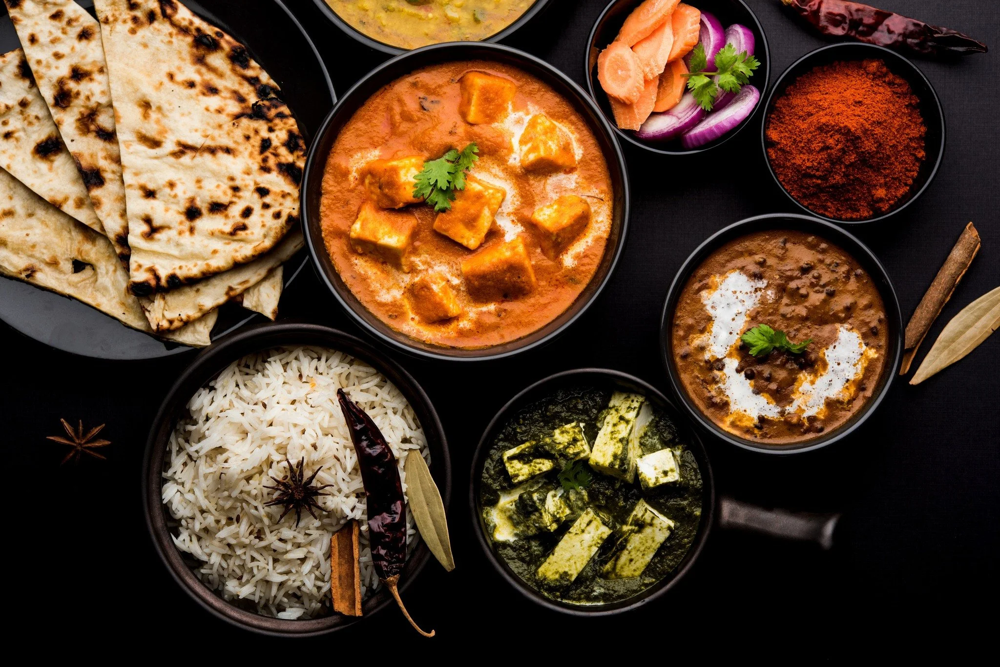 Assorted Indian dishes including naan, rice, paneer curry, lentil curry, spinach with cheese, and side condiments such as pickles, red chili powder, and dried chili peppers on a black background.