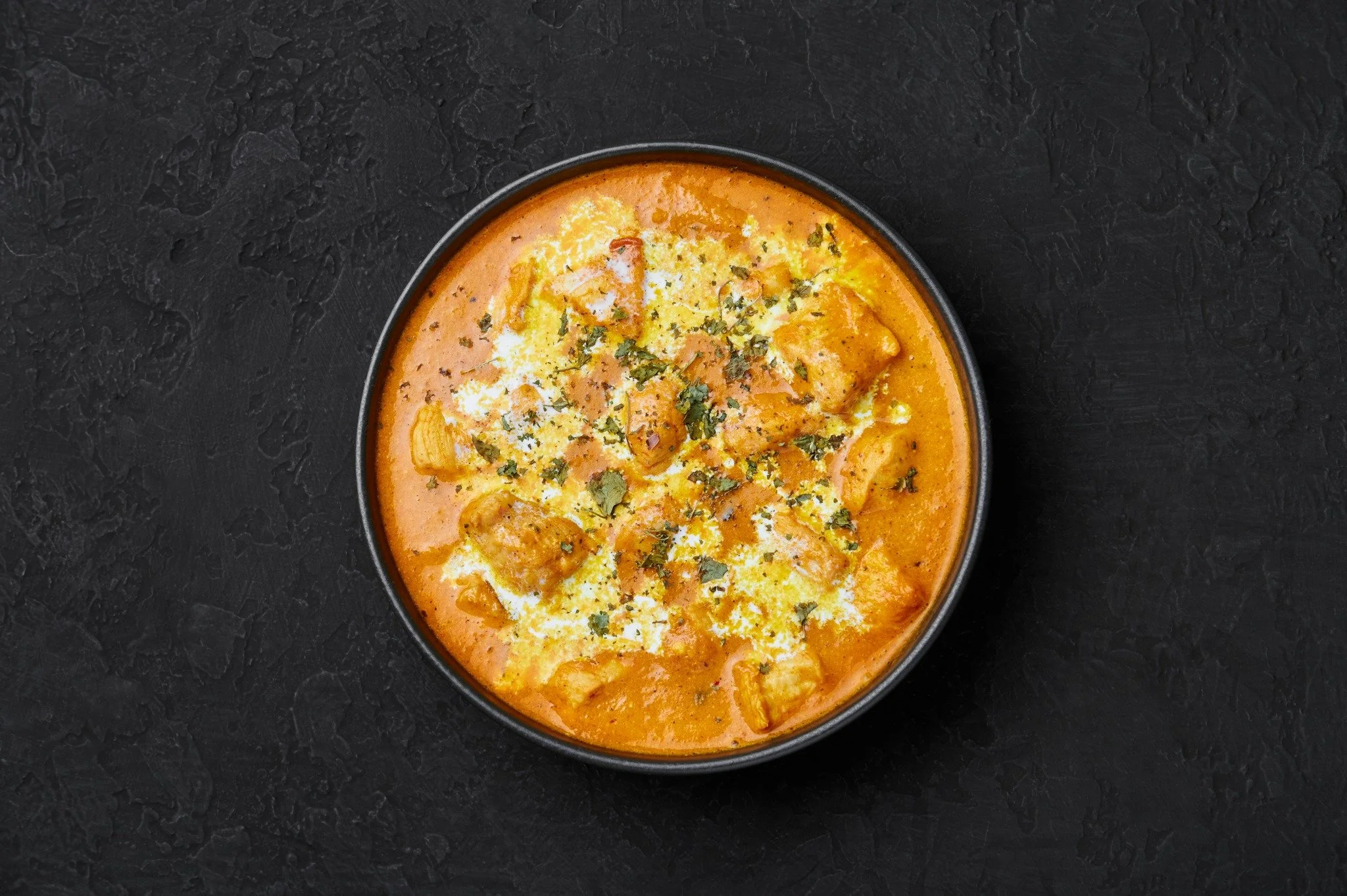 A bowl of Indian butter chicken curry topped with cilantro and cheese, on a black textured surface.