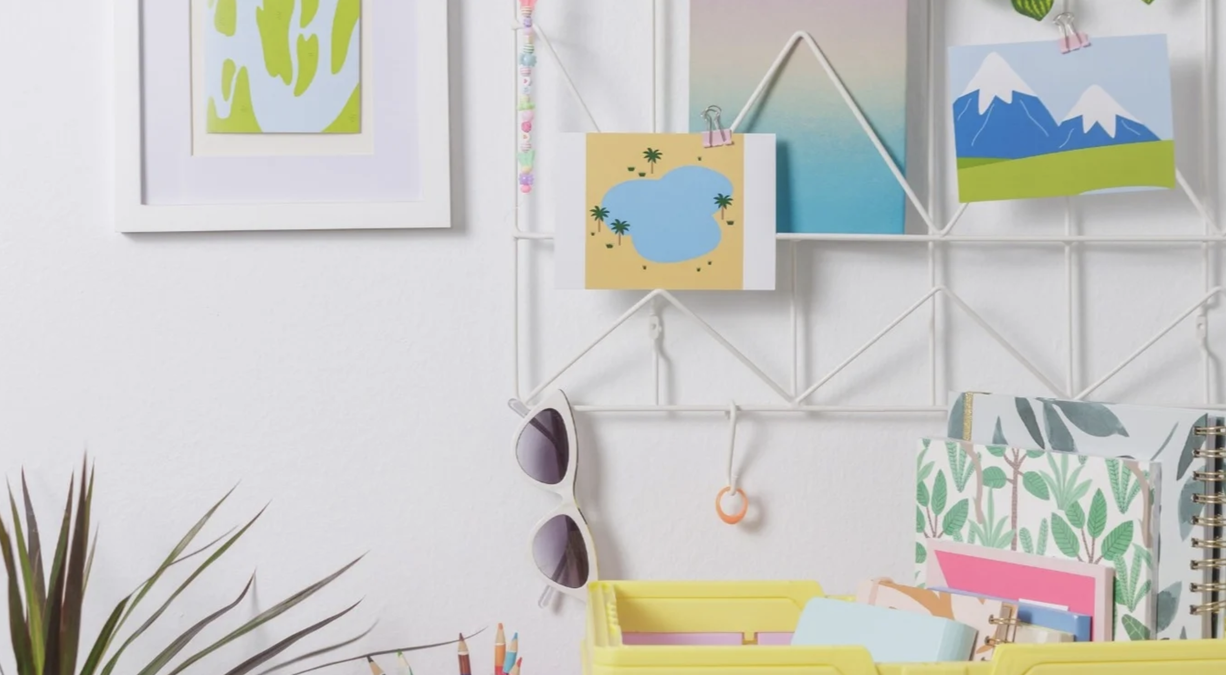 Decorative wall with framed and printed artwork, a white wire grid with hanging photos and accessories, sunglasses, books, and a yellow storage bin with school supplies, next to a plant.