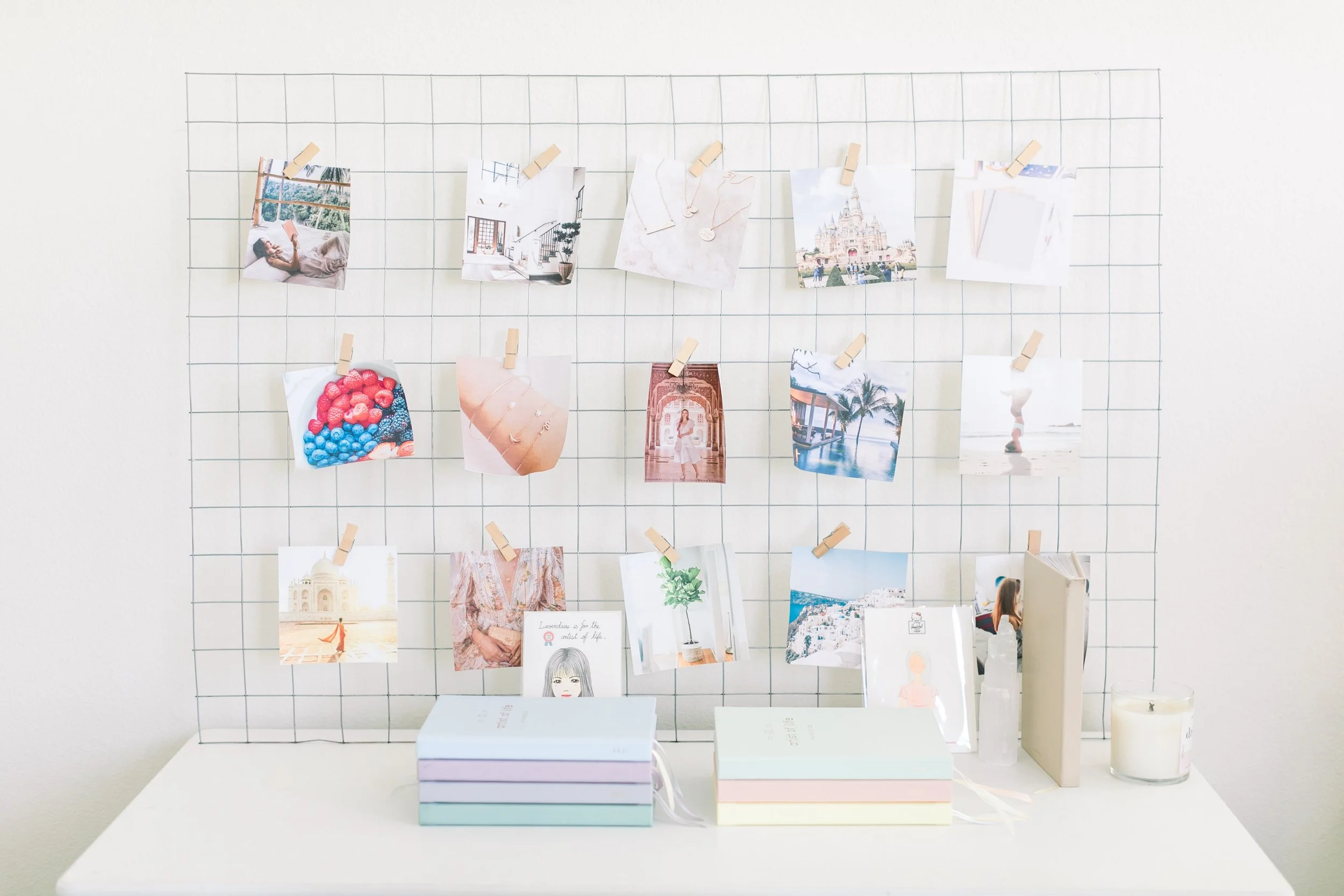 A wire grid wall with 15 photos attached by clothespins, positioned over a white table with books, a candle, and decorative items.
