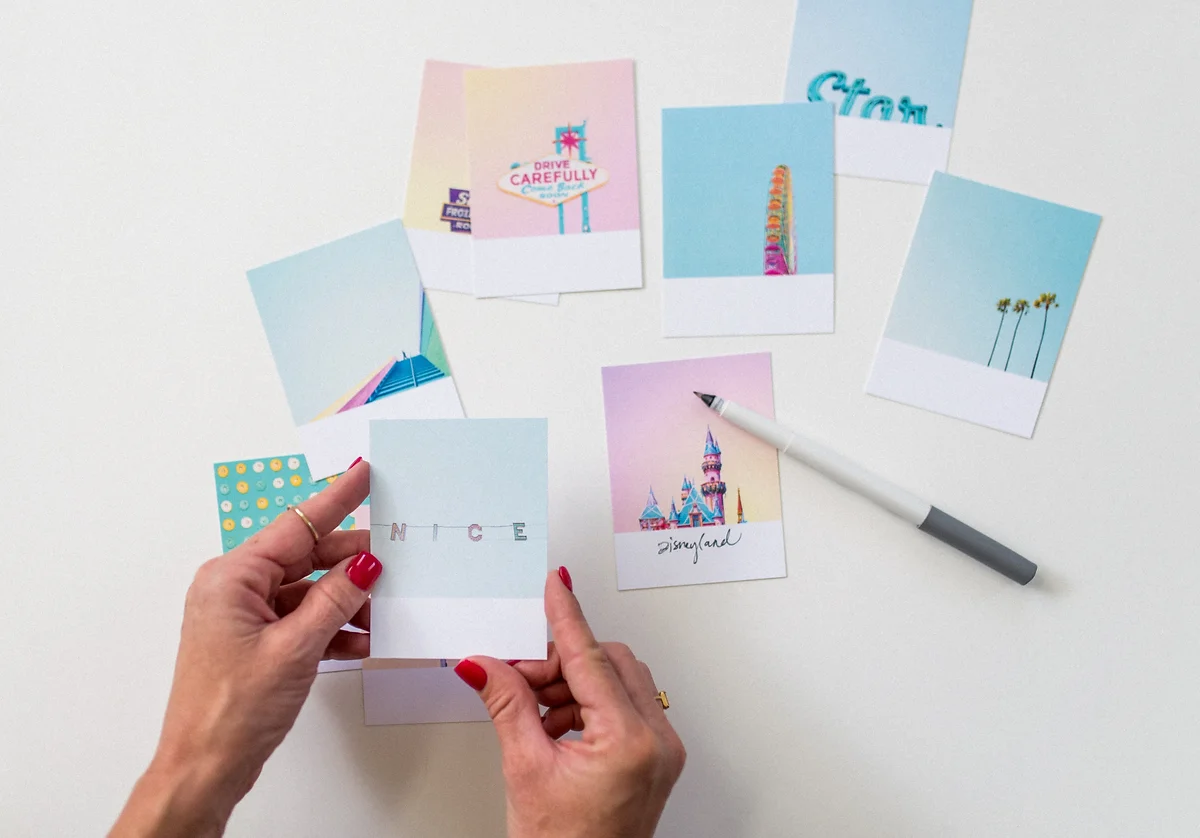 A person with red-painted nails holds a small photo card that spells out "NICE" with stitched letters. The person is sitting at a white table with various other colorful photo cards and a gray marker, some with images of Disneyland attractions, decor, and scenic views.