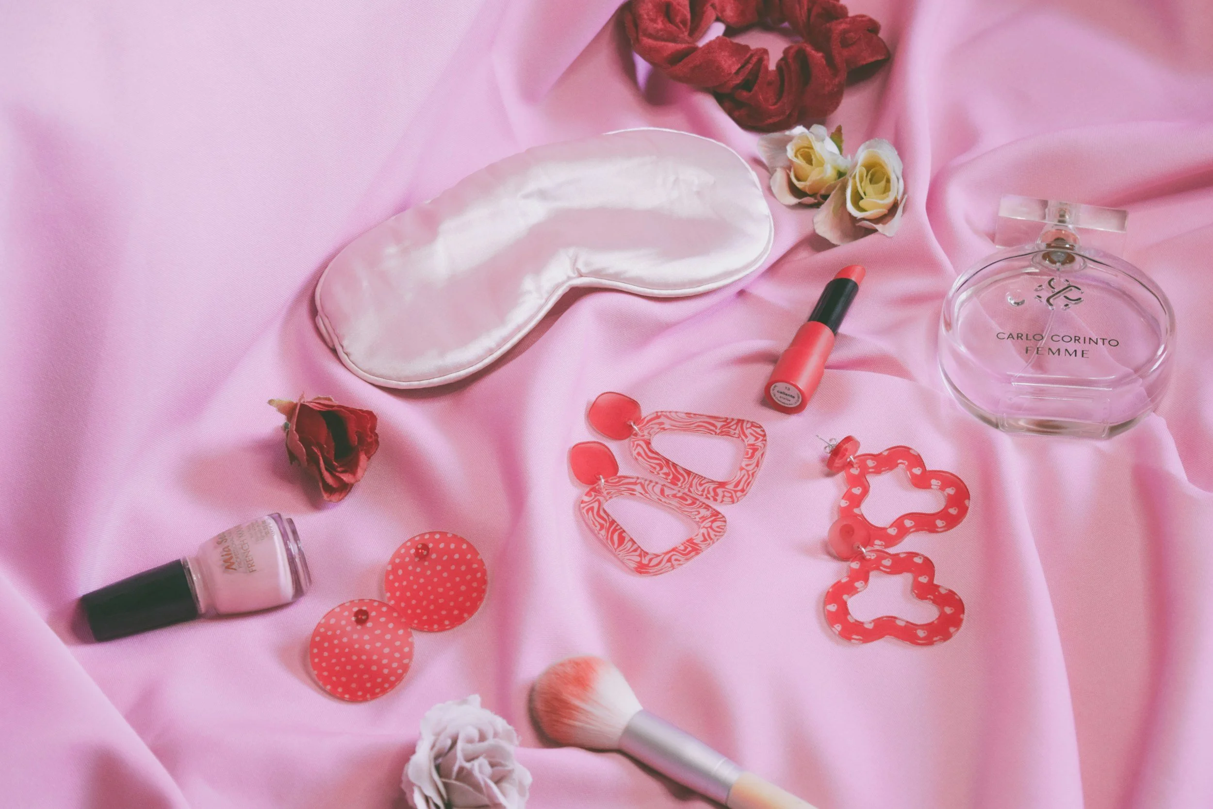Flat lay of pink and red accessories and beauty products on pink satin fabric, including a sleep mask, lipstick, perfume, polka-dot earrings, scrunchies, flowers, and a makeup brush.