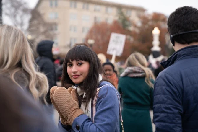 Young woman with dark hair wearing a blue coat and mittens at a protest or rally, surrounded by other people holding signs outdoors.
