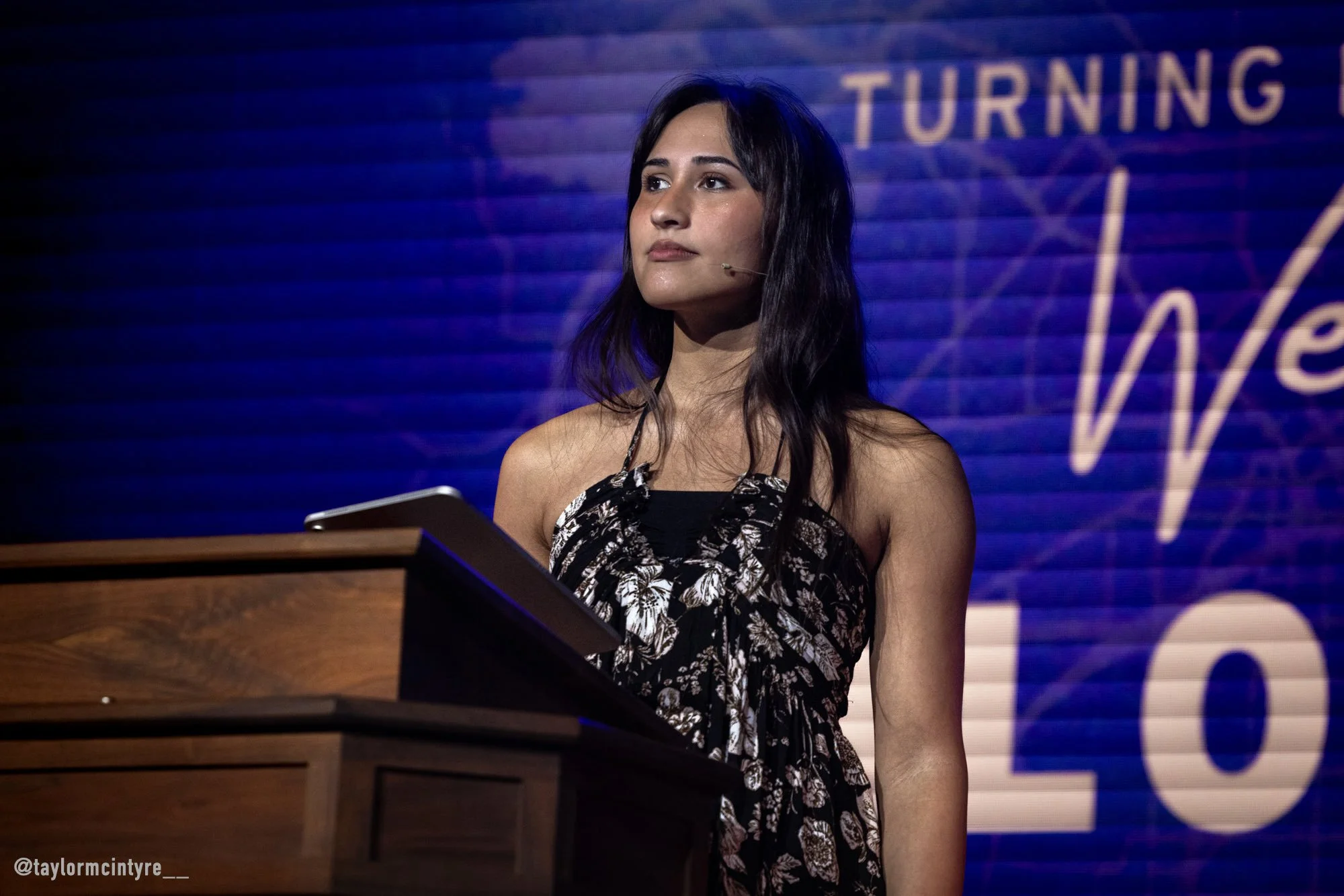 Woman at a podium with a microphone, standing in front of a blue background with partially visible text and logo.