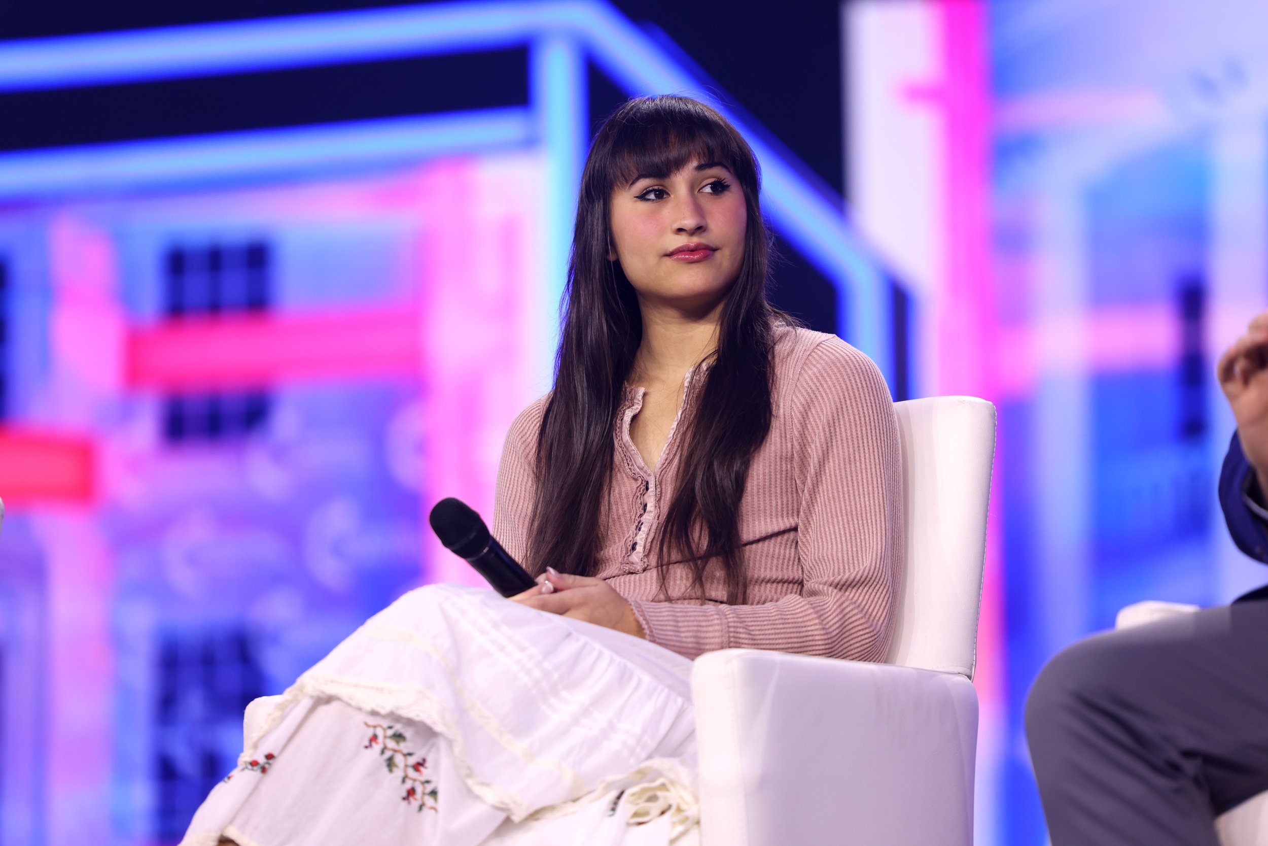 A young woman with long dark hair and bangs, wearing a pinkish-beige top and a white embroidered skirt, is seated on a white chair, holding a microphone, on a colorful stage with blue and pink lighting.