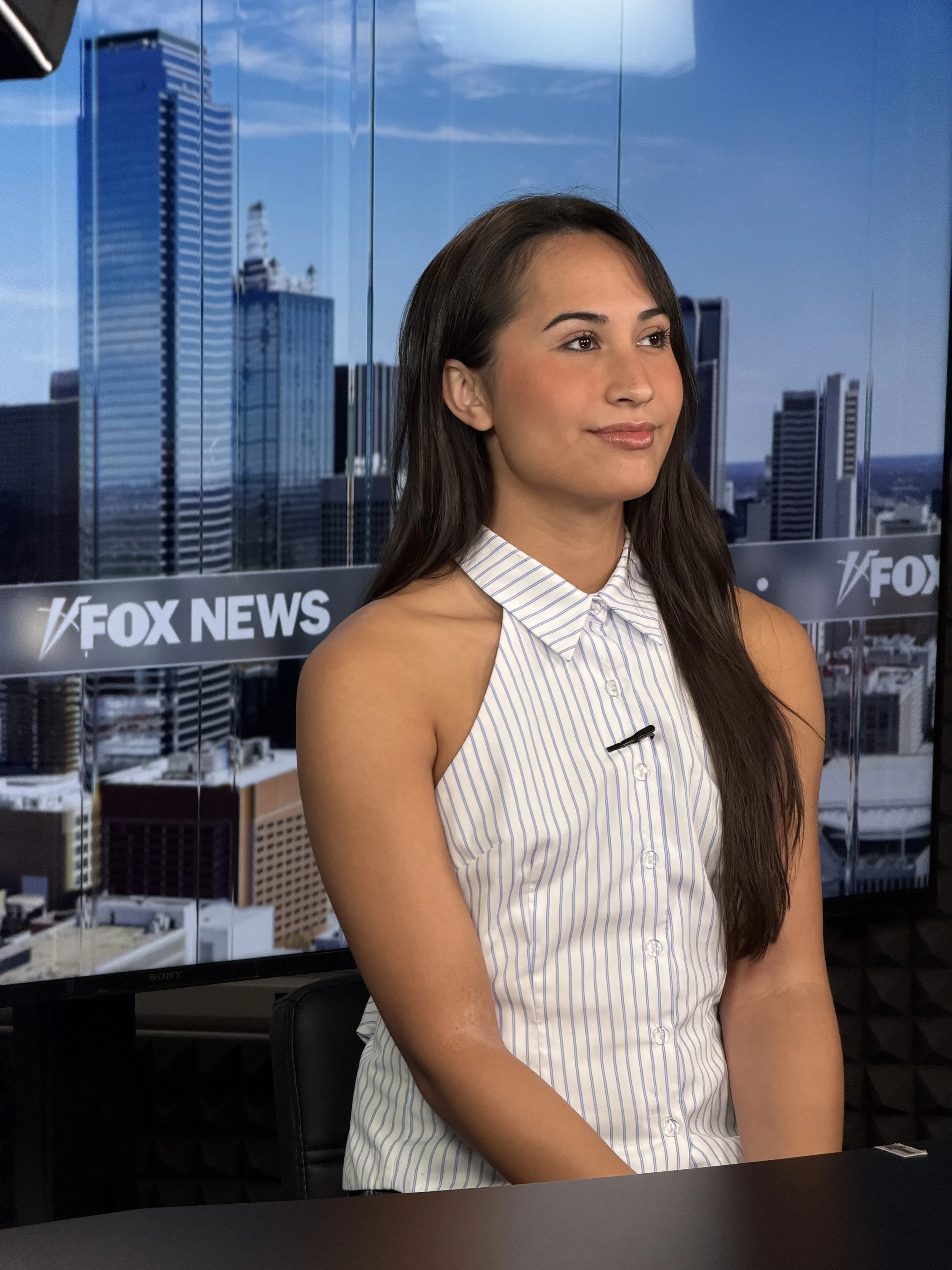 A woman with long dark hair, wearing a sleeveless white and blue striped shirt, sitting in front of a FOX News backdrop with city buildings in the background.