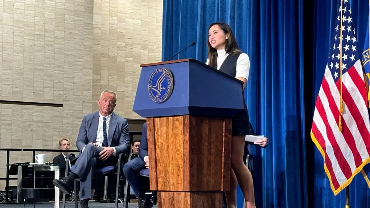 A woman is speaking at a podium on a stage with blue curtains and American flags, while a man in a gray suit sits and listens, with other men seated in the background.