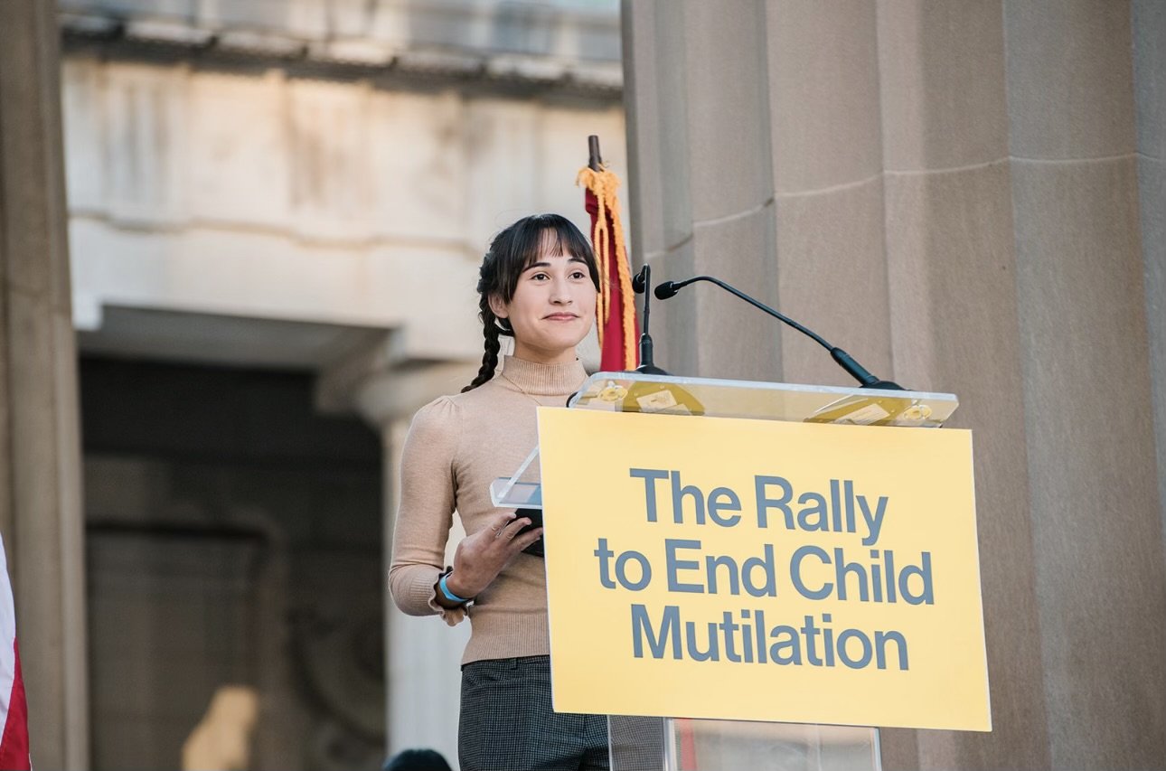 Young woman speaking at a rally with a yellow sign that reads 'The Rally to End Child Mutilation'.