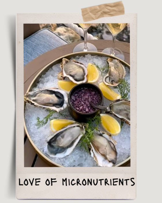 A round tray of fresh oysters on ice garnished with lemon wedges and herbs, with a small bowl of red onion or shallots in the center, two glasses of white wine in the background, and a cloth napkin on top of the photo.