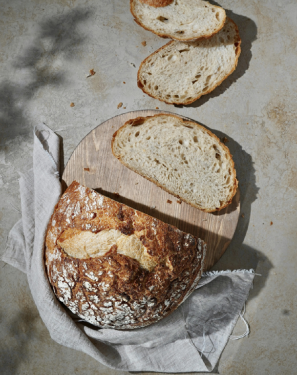 A loaf of rustic bread with a floured crust and a few slices cut, placed on a round wooden cutting board. The scene includes a white linen cloth underneath the bread and slices, on a neutral-colored surface.