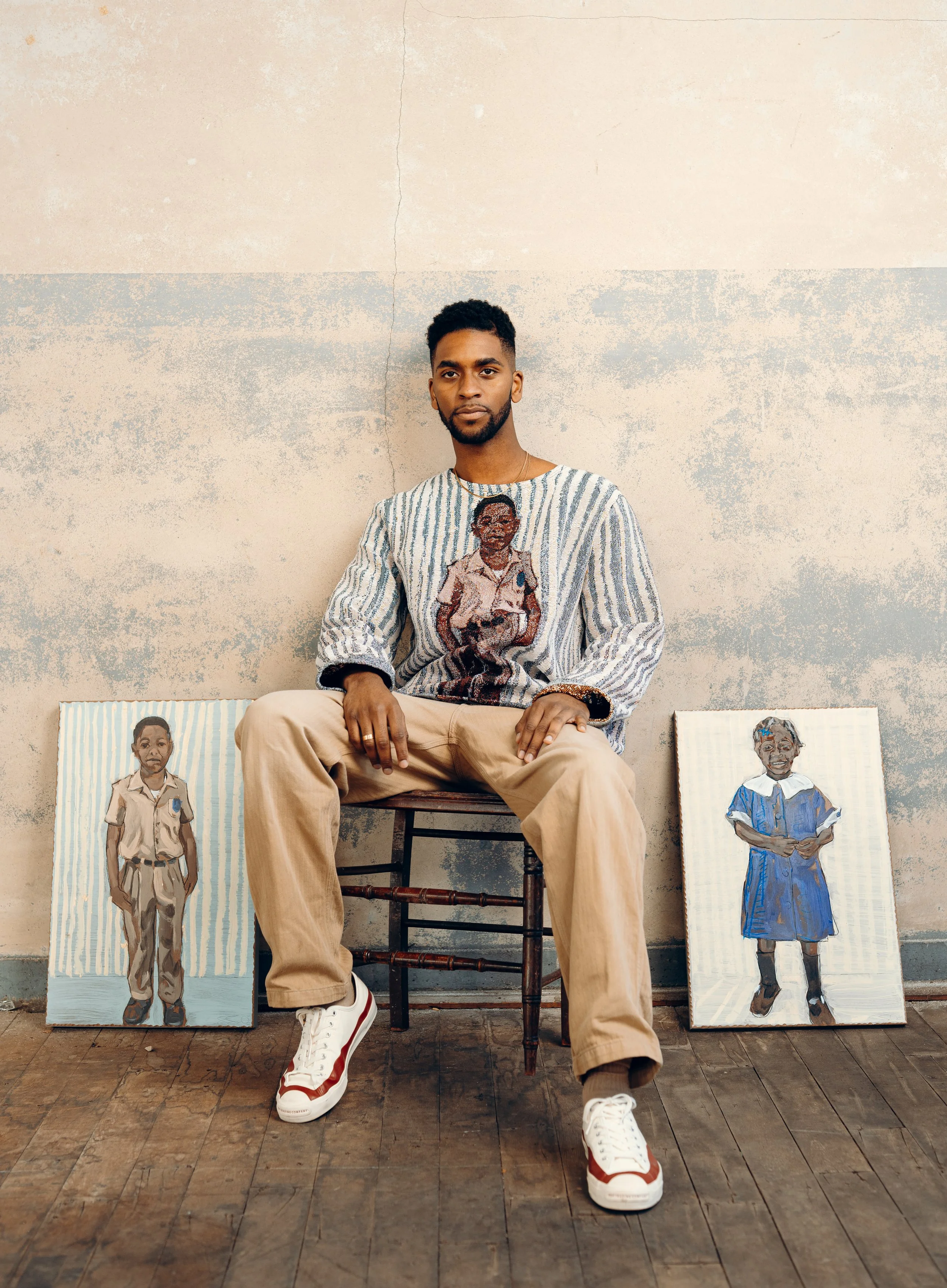 Un hombre sentado en una silla frente a una pared pintada, con dos retratos infantiles en el suelo a su lado.