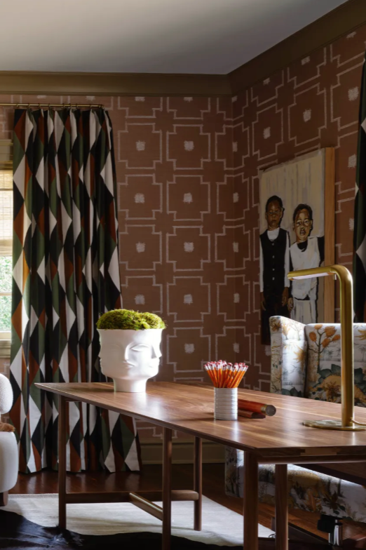 Una habitación con papel pintado marrón estampado, una mesa de madera con una maceta blanca con forma de cara llena de musgo verde, un recipiente con lápices rojos y un grifo dorado. En la pared hay un gran cuadro en el que aparecen dos niños, y junto a la ventana hay unas cortinas geométricas de varios colores.