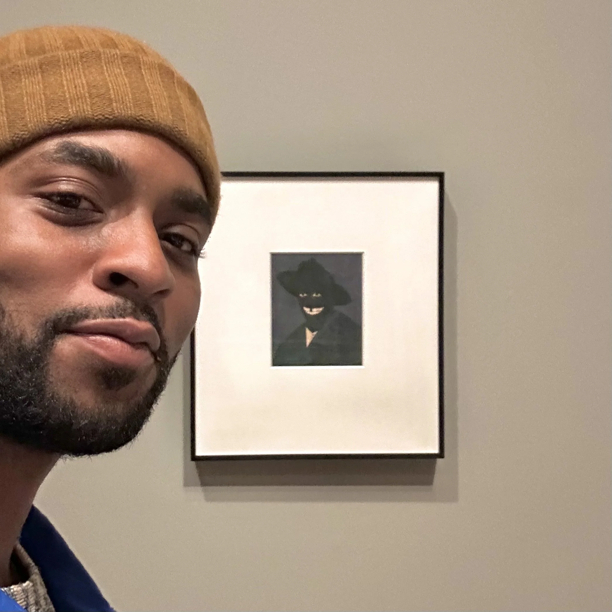 Un hombre con un gorro marrón y una chaqueta oscura se hace un selfi delante de un cuadro que representa a una figura con un sombrero de ala ancha y una sonrisa pícara.