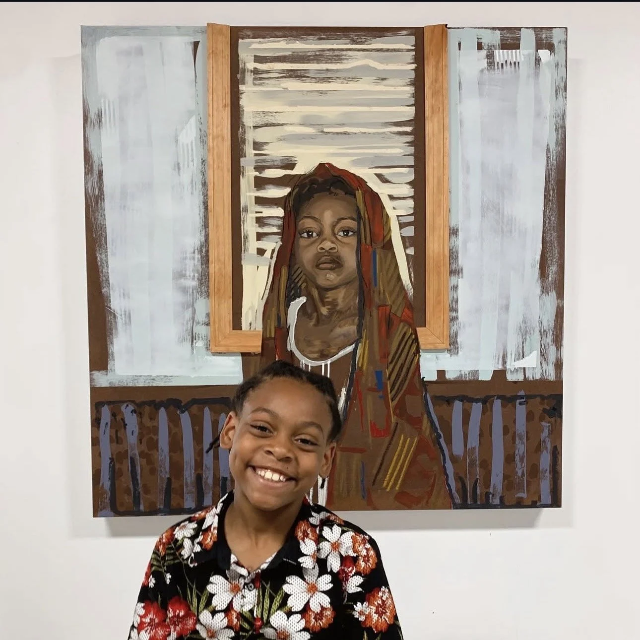 Una joven sonriente, con el pelo trenzado y una camisa de flores, posa delante de un colorido retrato de una mujer con un pañuelo en la cabeza, con unas contraventanas y una pared pintadas como fondo.