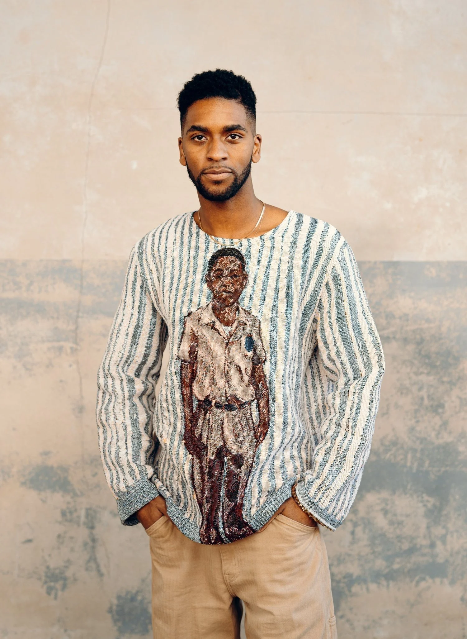 Un joven de pelo oscuro y barba posa de pie frente a una pared lisa; lleva un jersey a rayas con un gran estampado gráfico de un hombre uniformado en la parte delantera y pantalones de color caqui.