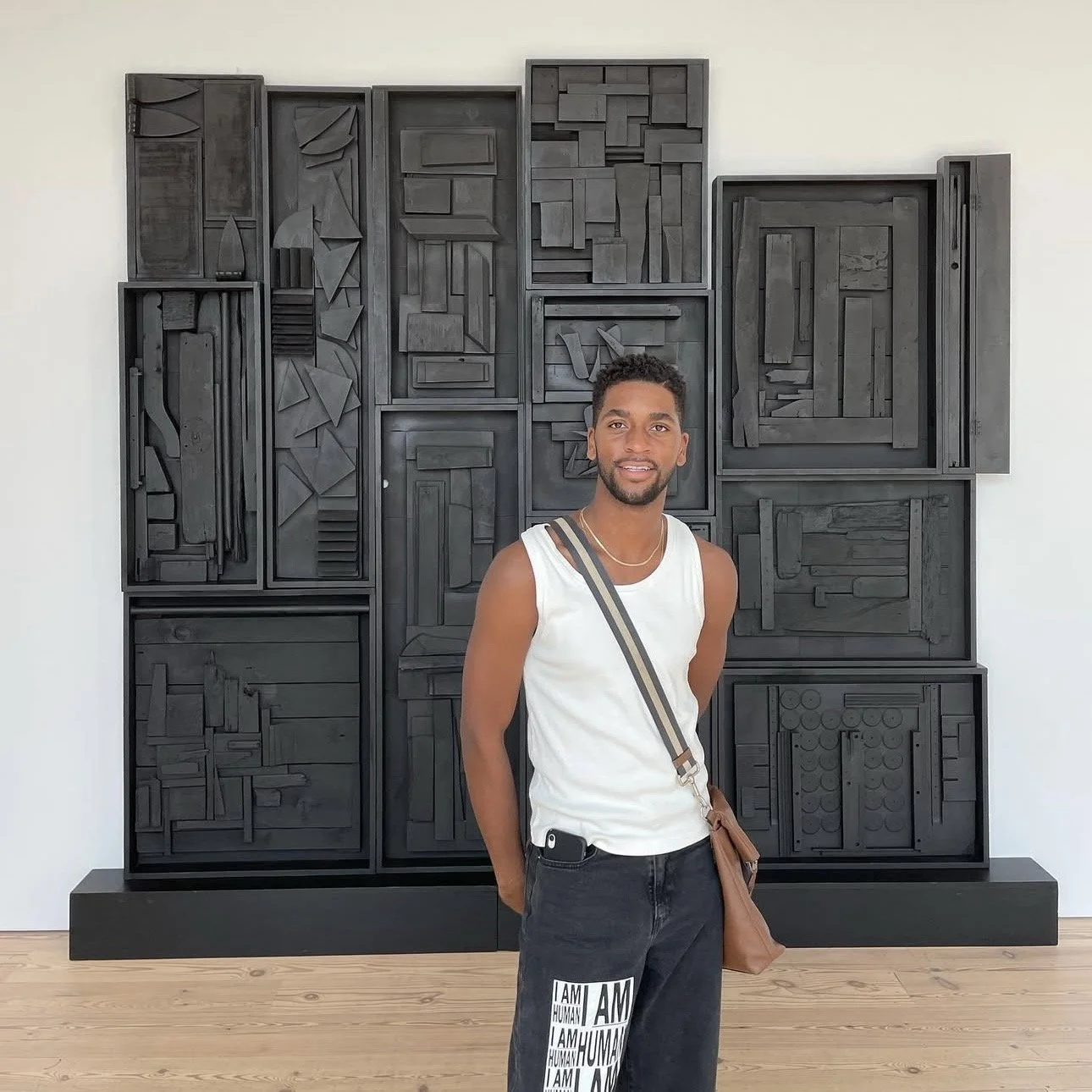 Un joven con el pelo rizado y barba, de pie frente a una escultura abstracta moderna de color negro sobre una pared blanca en una galería de arte. Lleva una camiseta blanca sin mangas, pantalones negros con letras blancas y un bolso de color beige con una correa cruzada sobre el pecho; lleva un teléfono en la cintura.